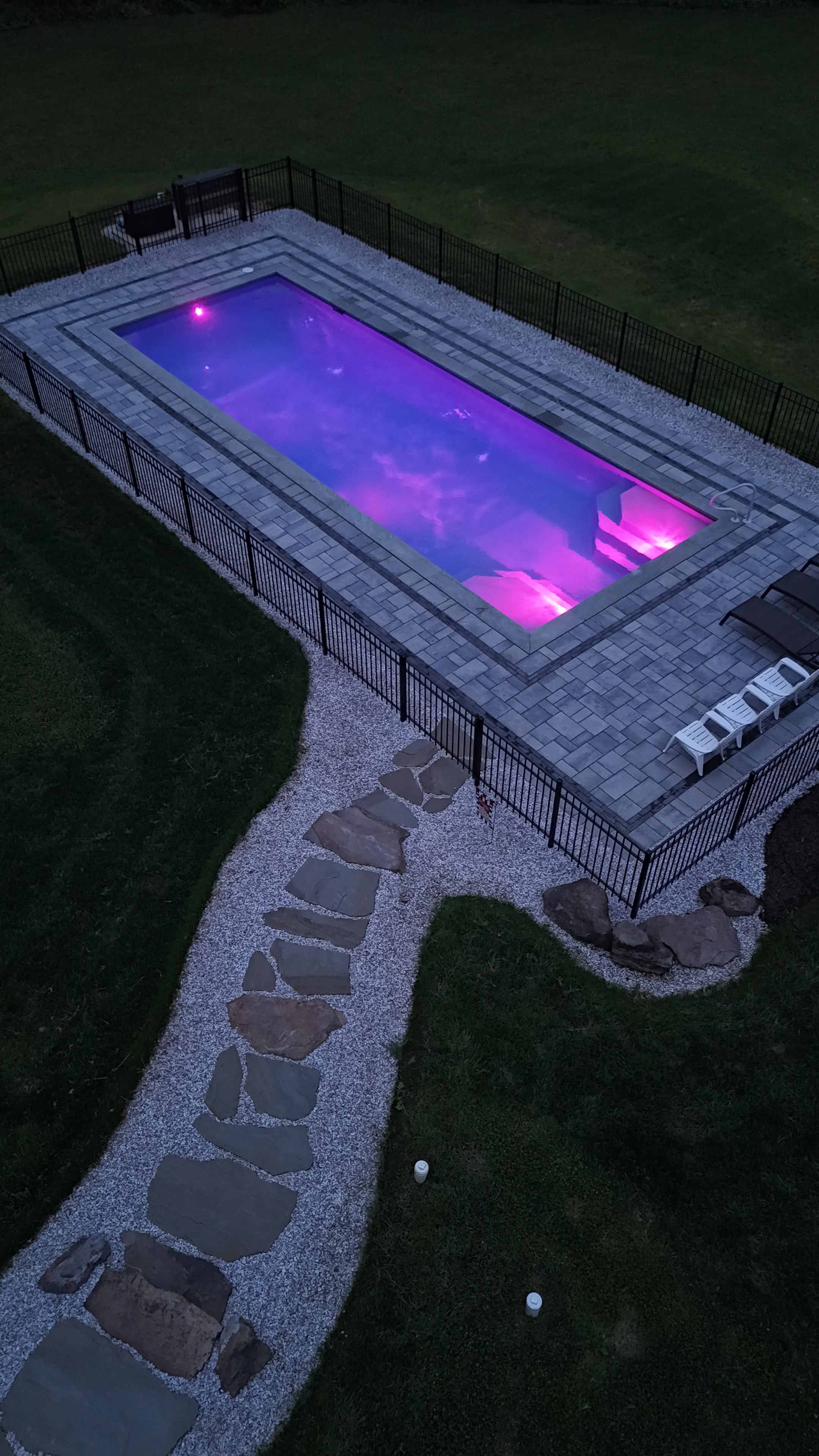 LandPro aerial night shot of pool glowing purple with paver surround and flagstone path leading to patio