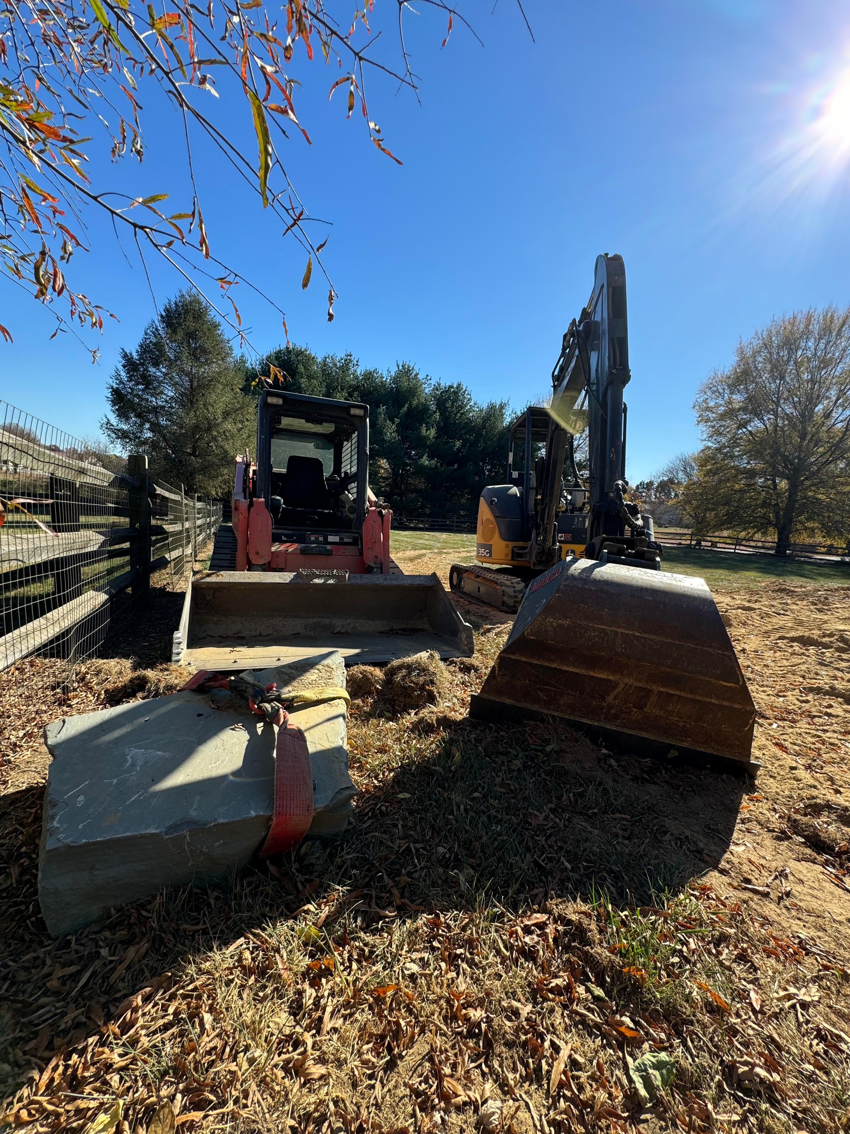 Under construction — skid steer and excavator on site with boulders staged for landscaping