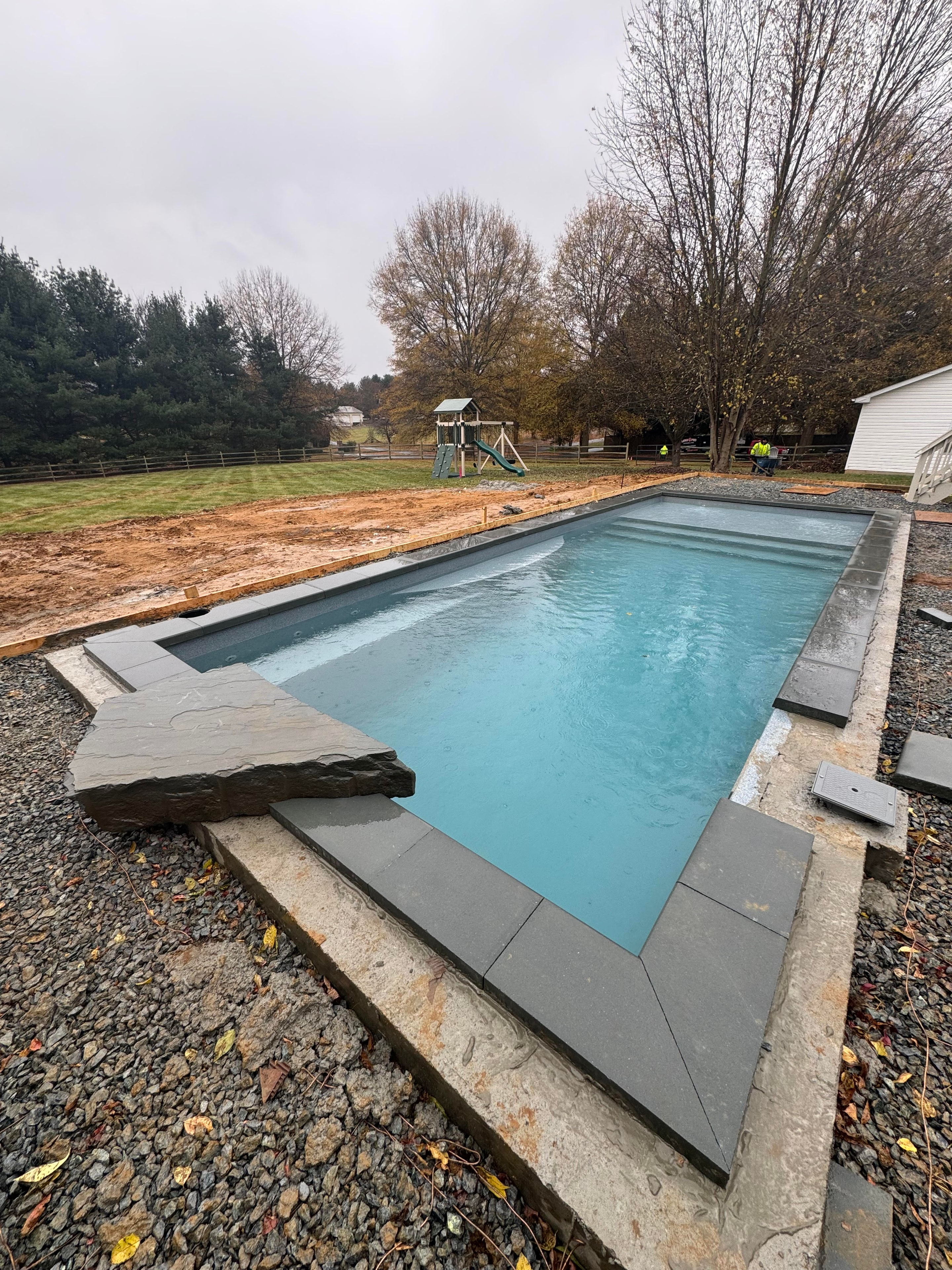 Under construction — pool shell installed with coping and gravel surround, playground visible in background