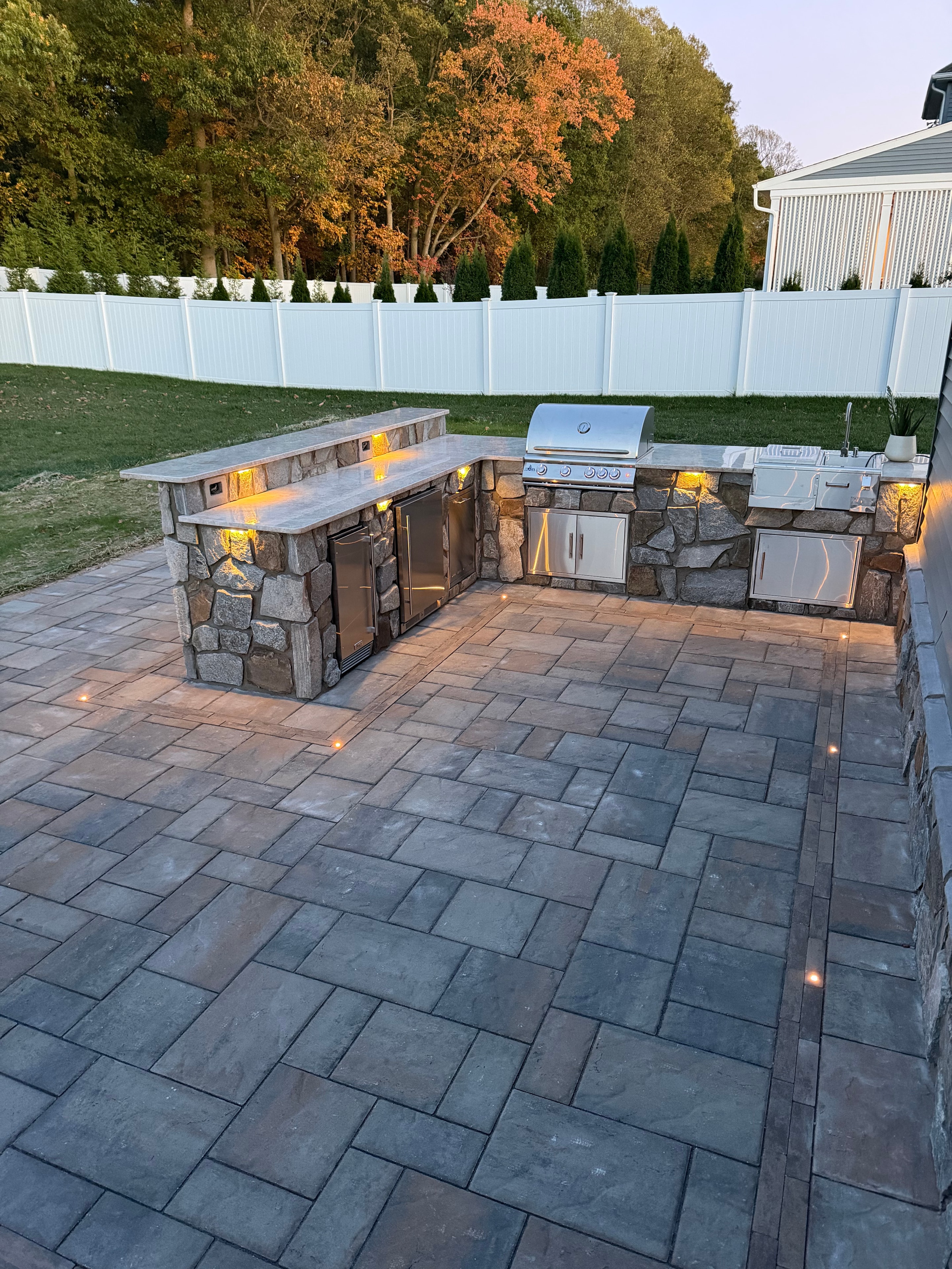 Chef's Dream — aerial dusk shot of crew member flying drone over paver patio with LED dot lights, outdoor kitchen with stone surround and granite countertop, composite deck alongside