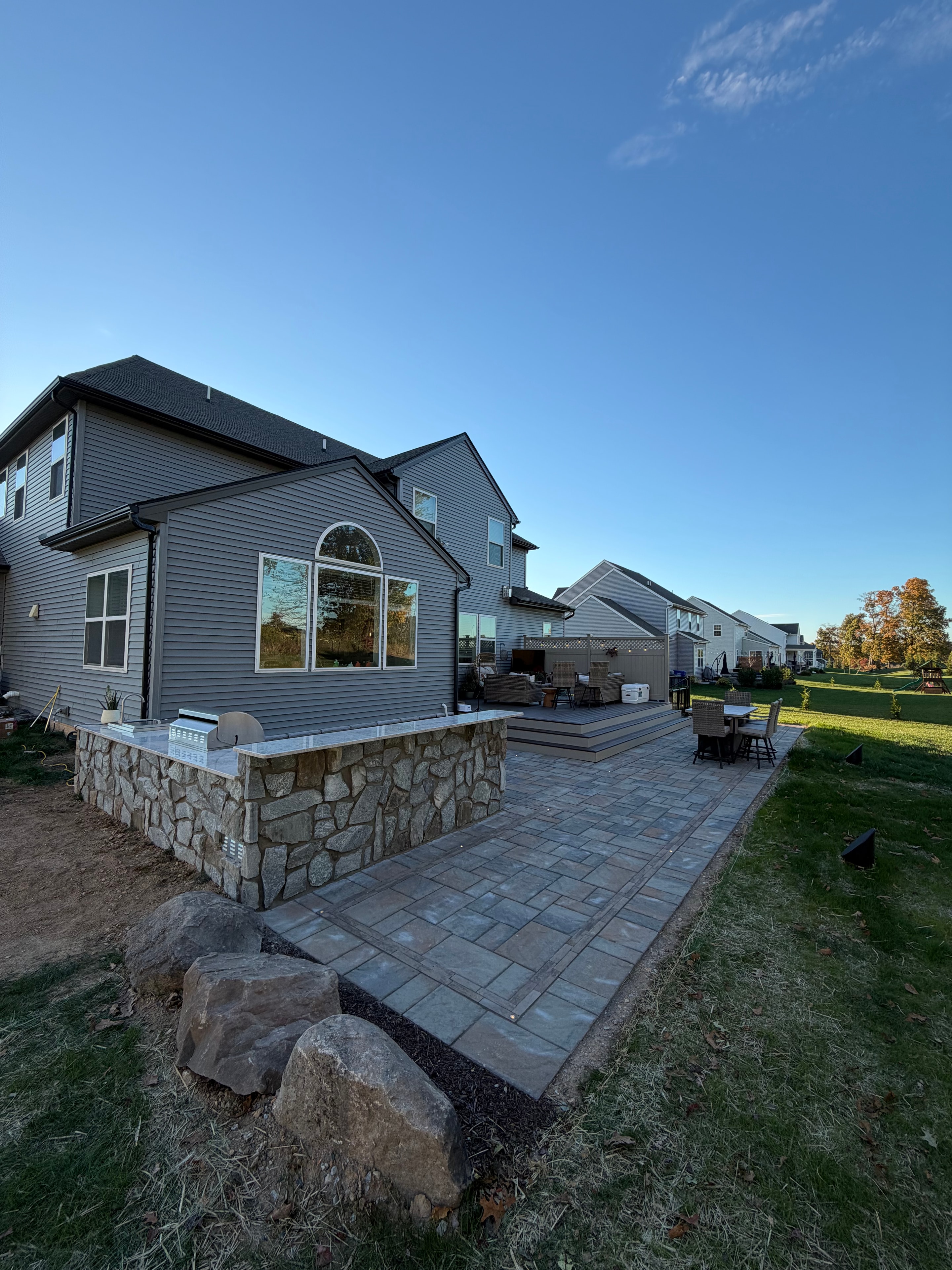 Chef's Dream — daytime wide shot of paver patio beside modern gray metal-sided house with stone retaining wall, boulder accent, and composite deck with pergola in background