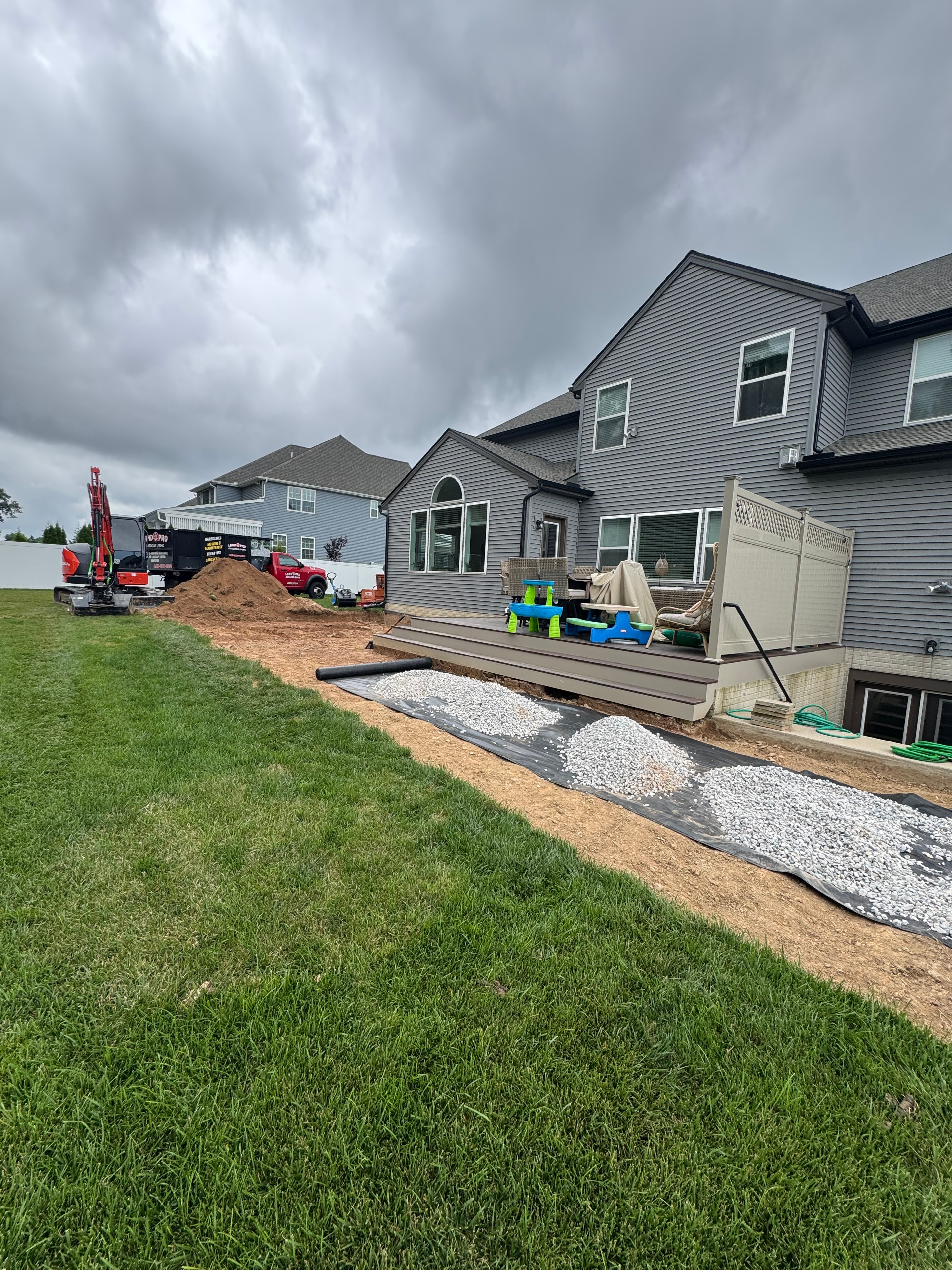 Chef's Dream — early site work showing composite deck beside gray metal-sided house, gravel base laid, red excavator and dumpster in background, overcast sky