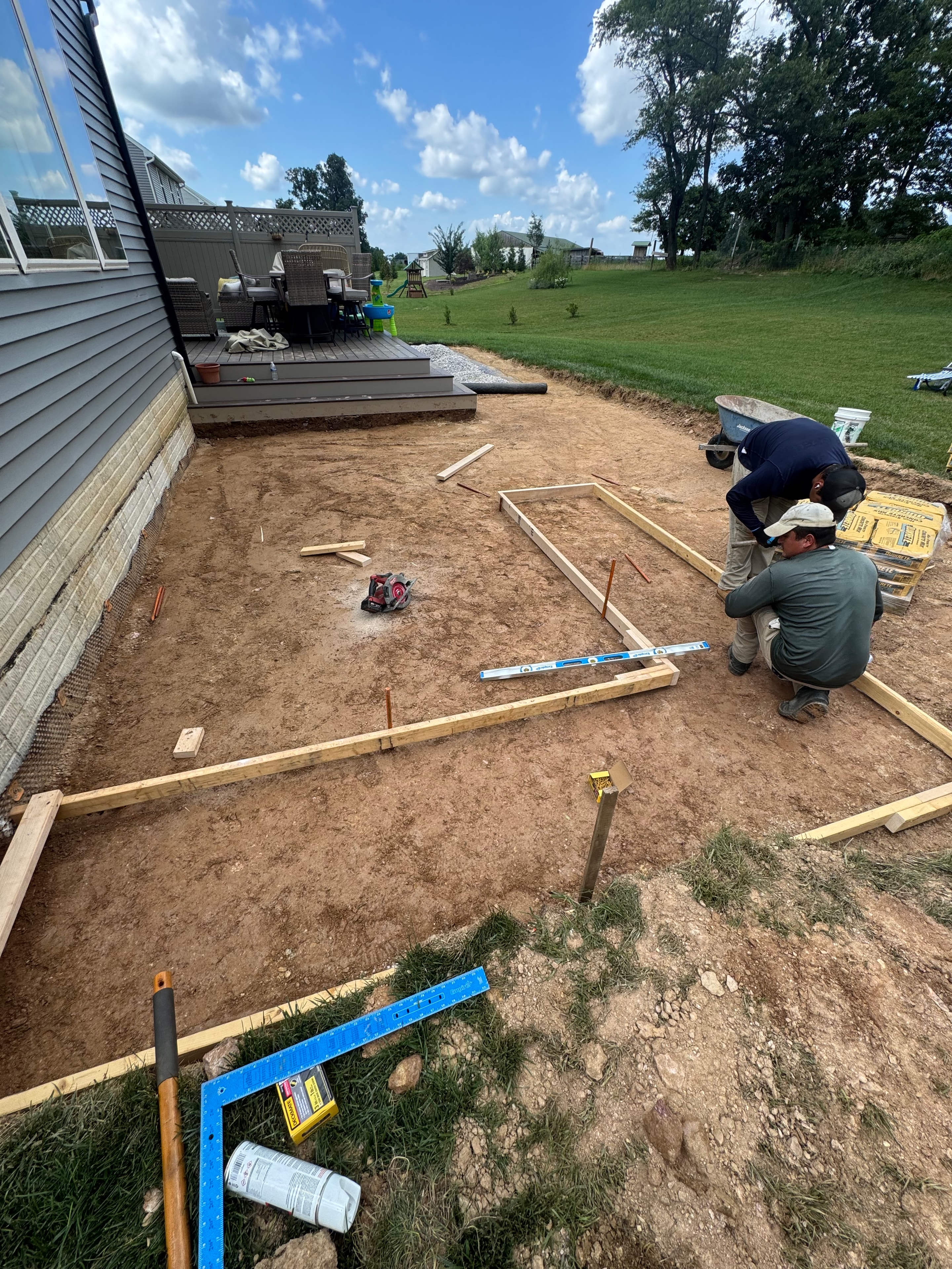 Chef's Dream — two crew members kneeling on bare dirt setting concrete forms for patio, lumber and tools spread out, composite deck visible in background, blue sky