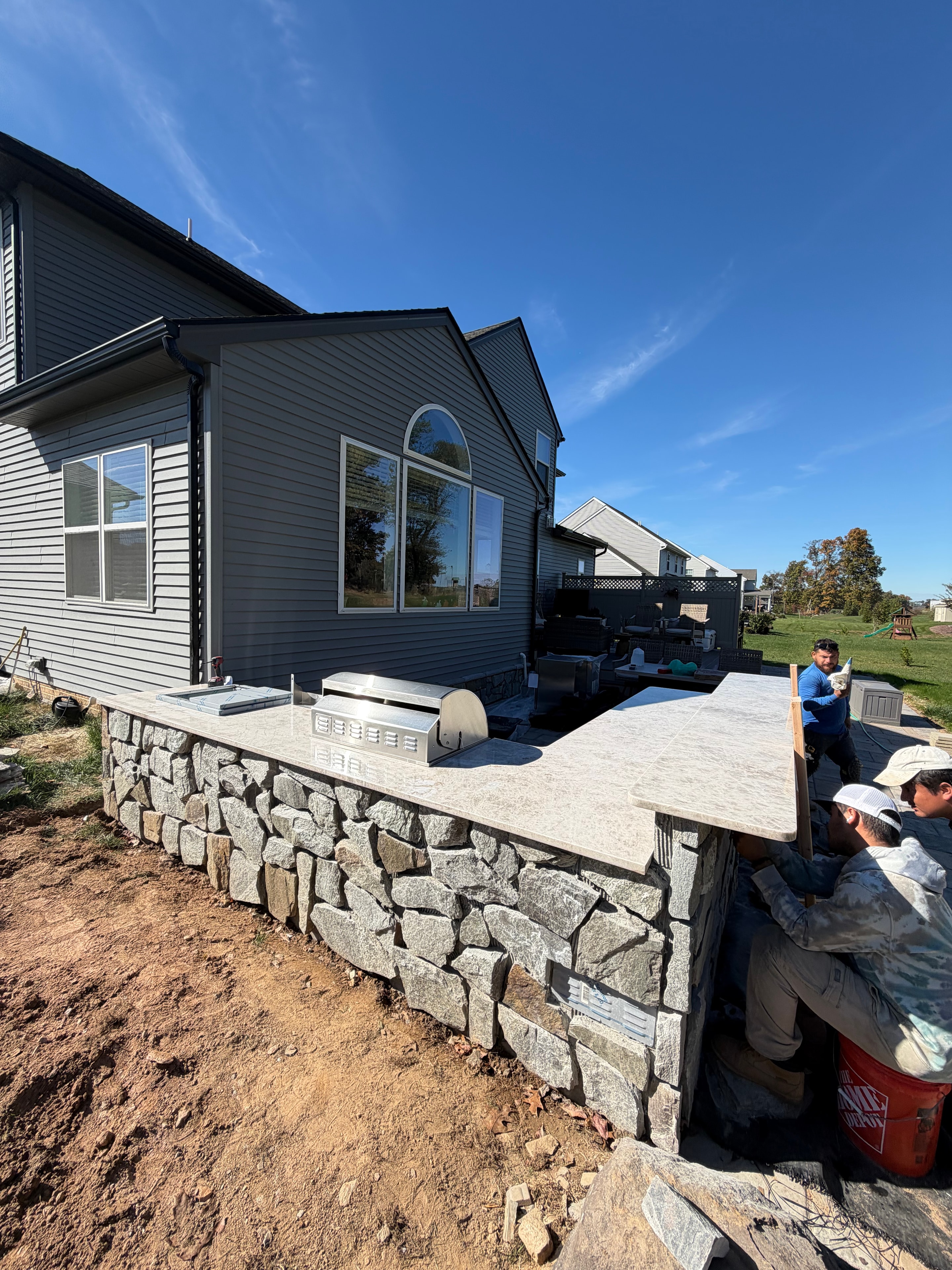 Chef's Dream — crew installing granite countertop on outdoor kitchen stone surround, grill set in place, blue sky and gray metal-sided house backdrop