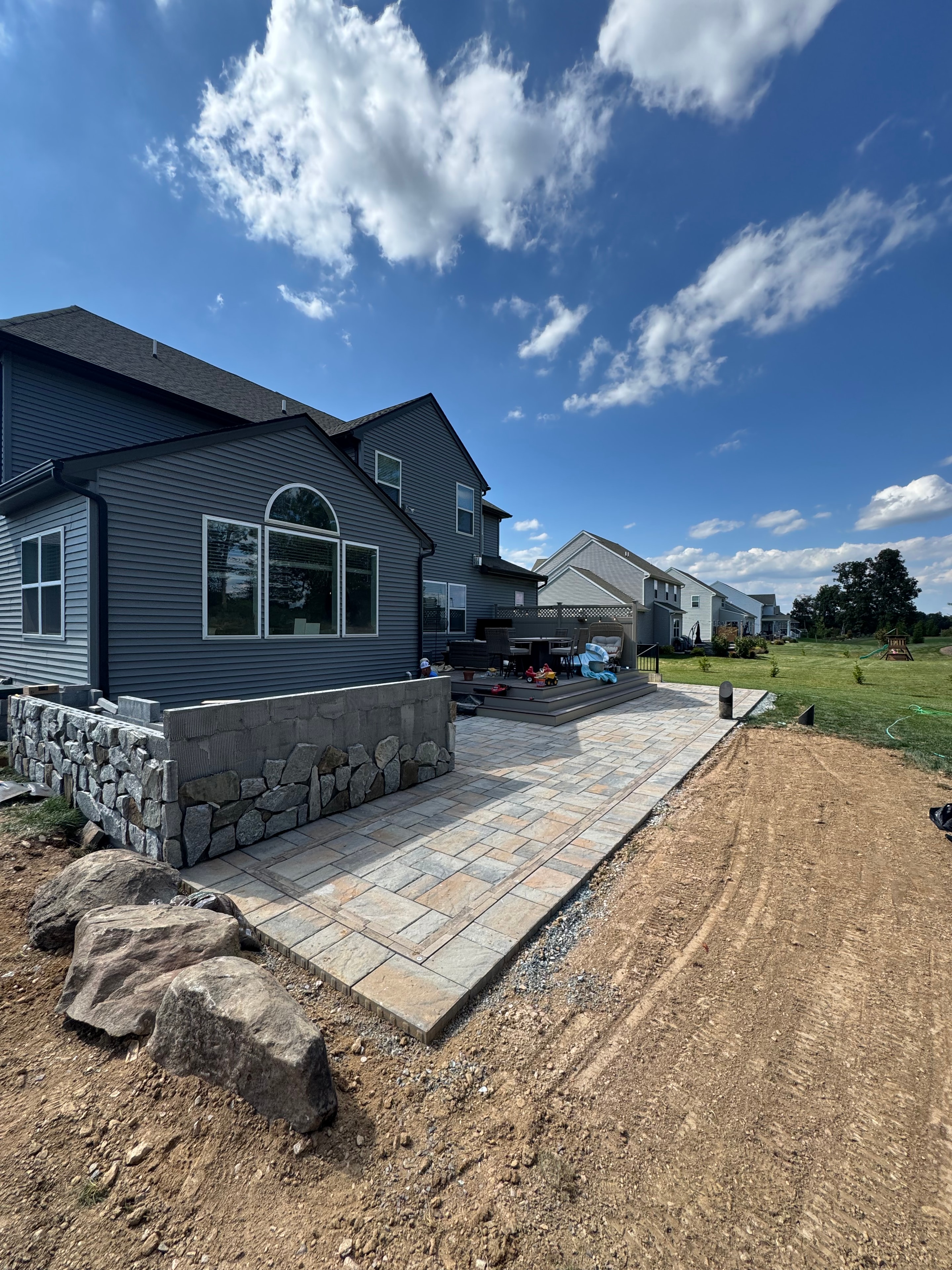 Chef's Dream — wide shot of outdoor kitchen stone surround and Techo Bloc patio taking shape, retaining wall with boulder base visible, crew and equipment on site, blue sky