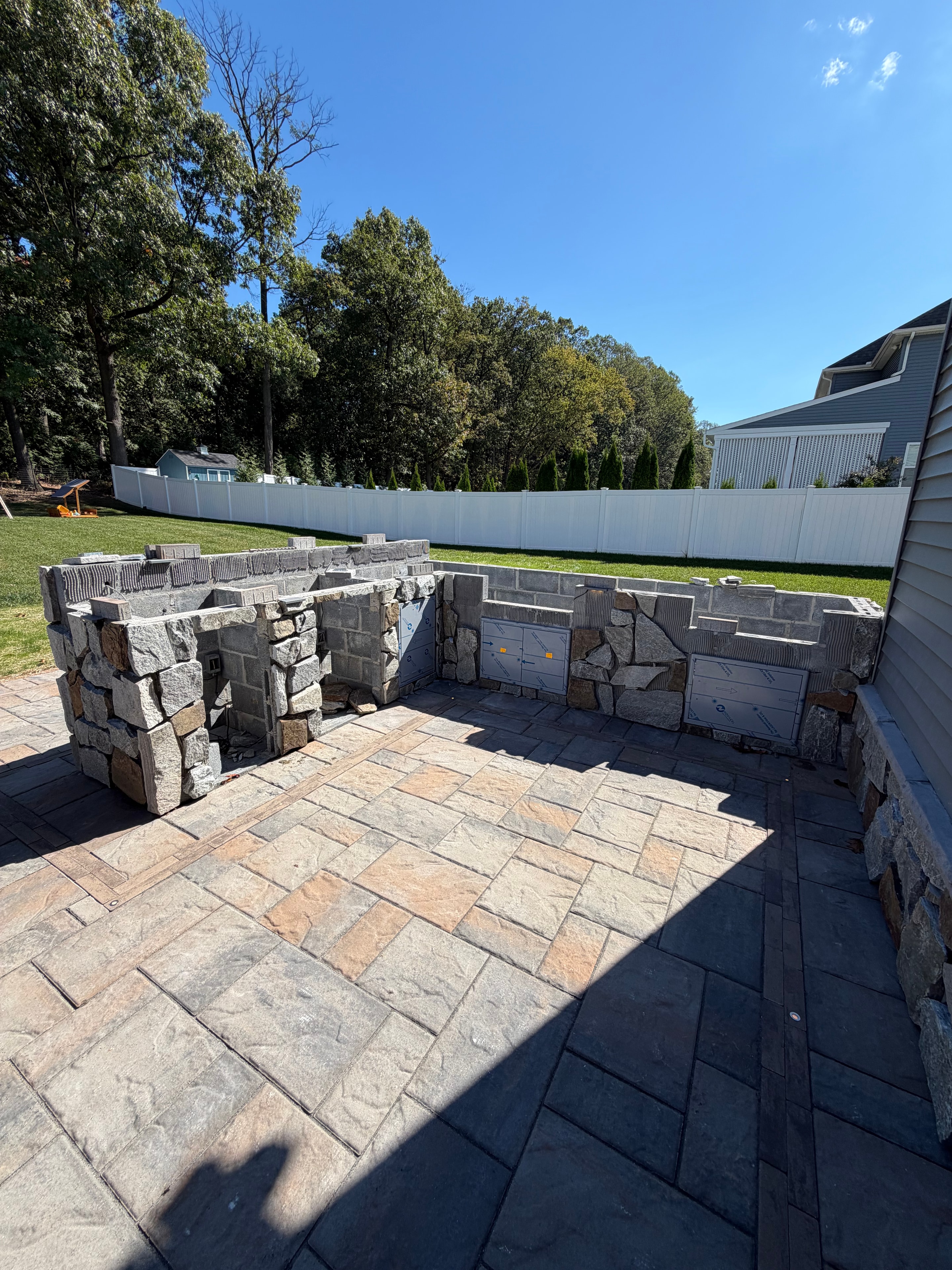 Chef's Dream — outdoor kitchen stone surround with stainless appliance cutouts framed in, Techo Bloc patio laid, green trees and blue sky backdrop