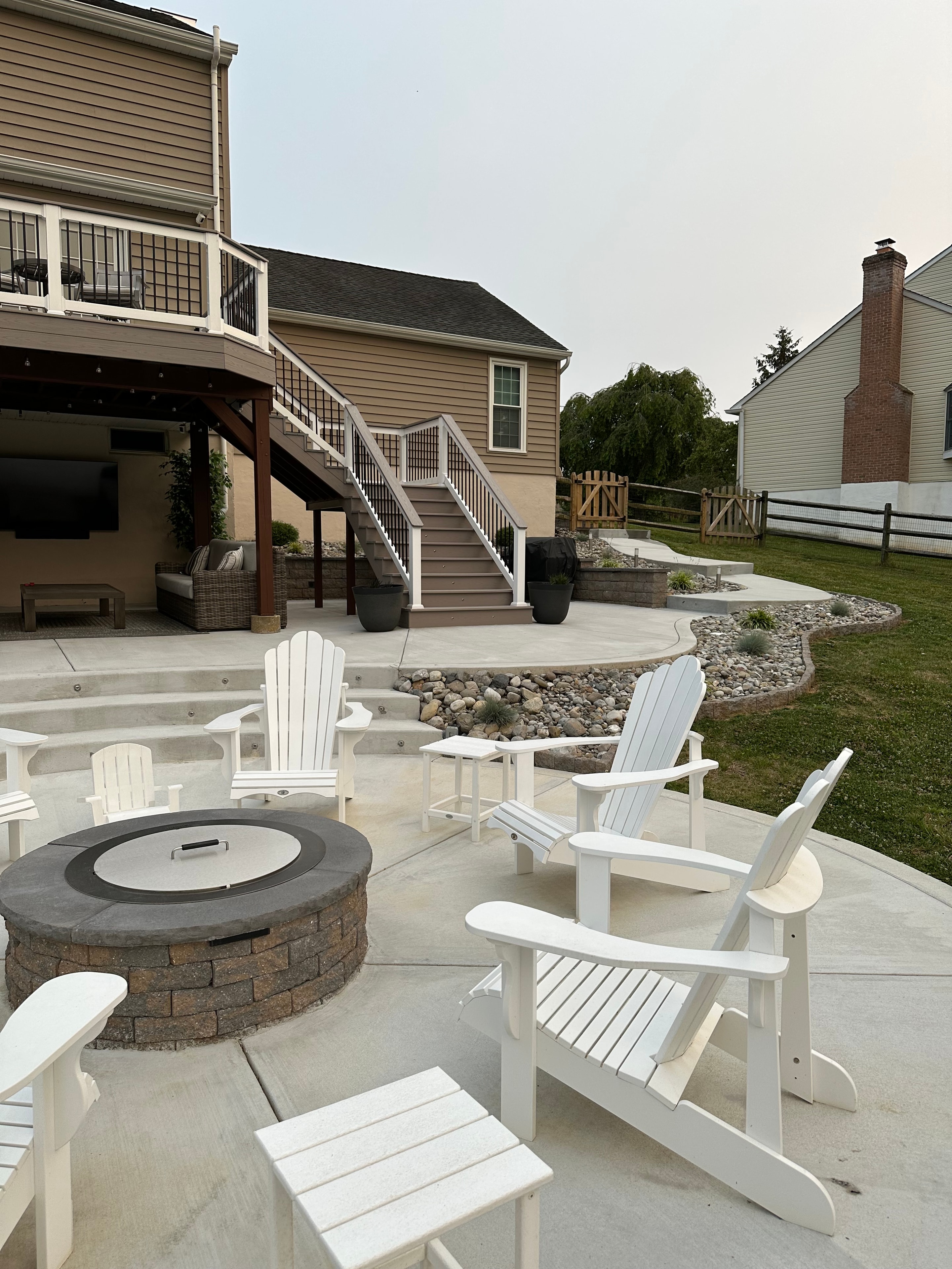 Concrete Jungle — daytime shot of circular fire pit seating area with white Adirondack chairs, concrete patio, multi-level deck staircase, and under-deck covered space