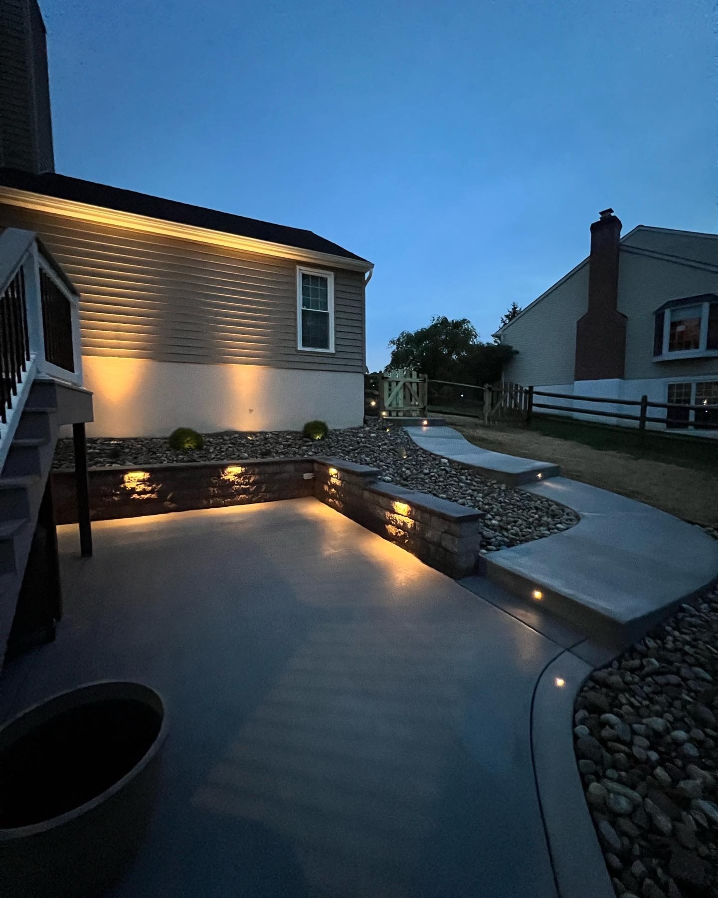 Concrete Jungle — dusk ground-level shot of concrete patio with LED-lit granite sitting wall, LED step lights leading to upper patio, and river rock landscaping