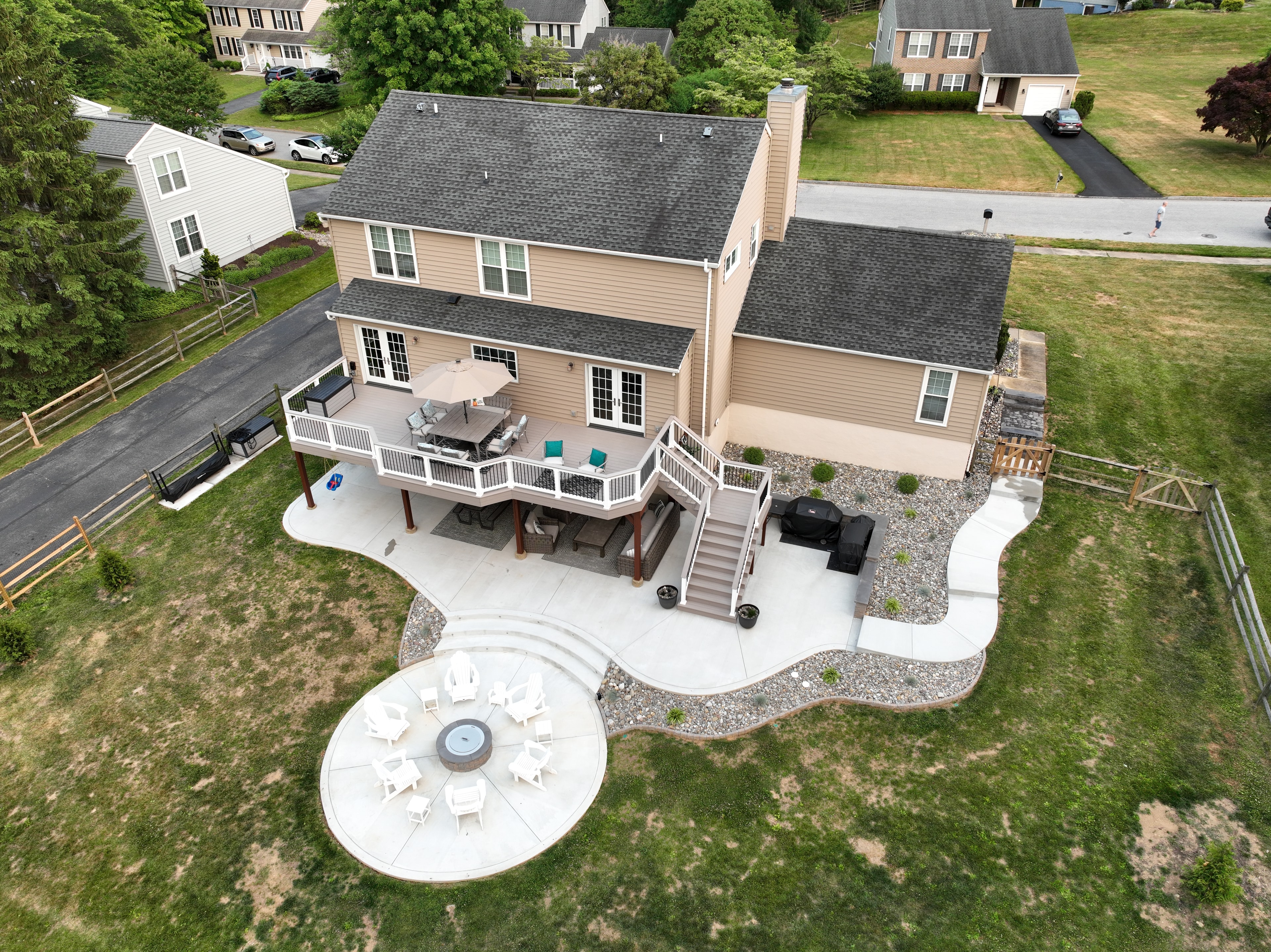 Concrete Jungle — aerial daytime shot of full backyard with freeform concrete patio, circular fire pit pad with white Adirondack chairs, multi-level deck, under-deck covered patio, and river rock landscaping