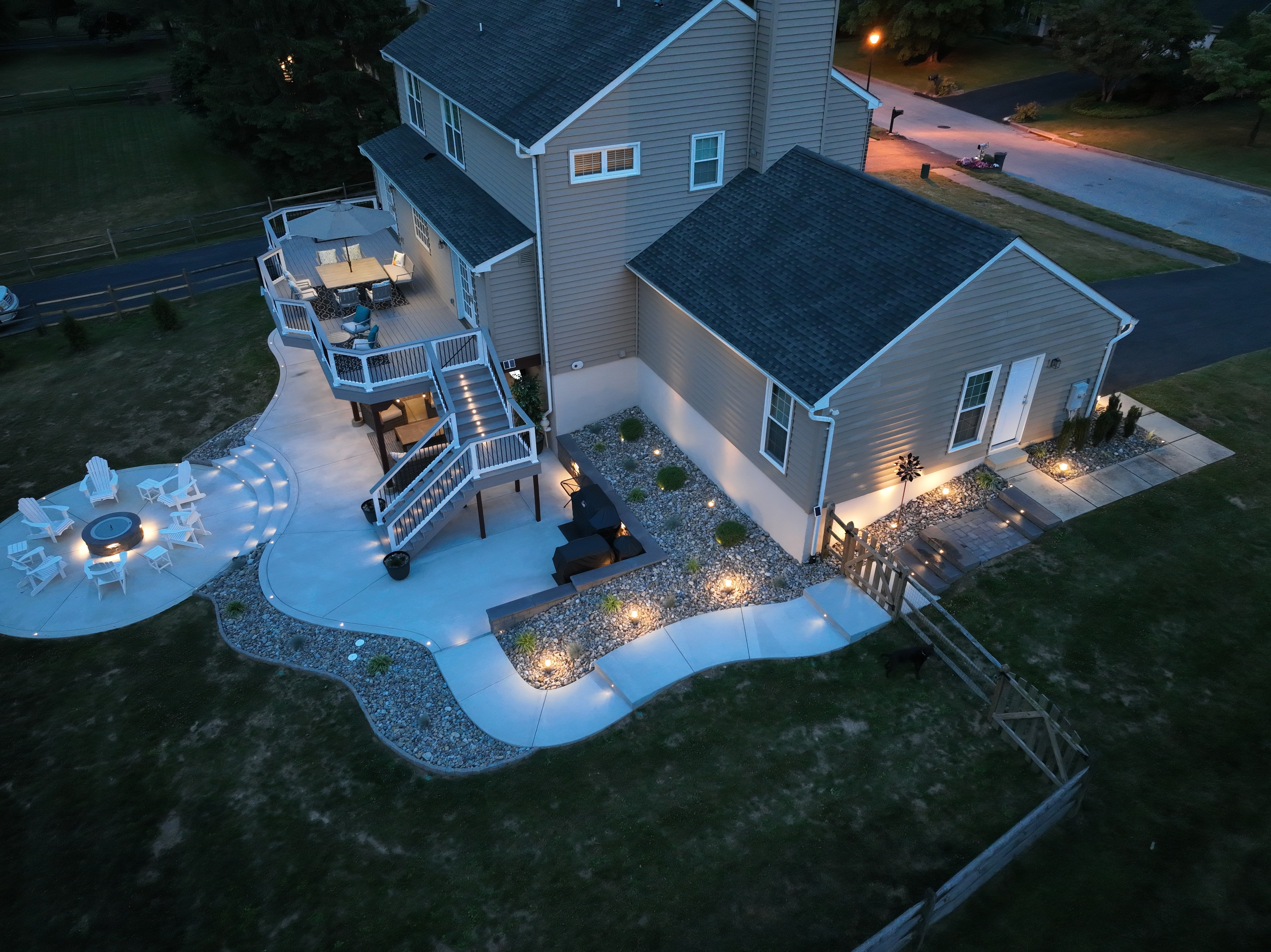 Concrete Jungle — aerial daytime angled shot showing freeform concrete patio, circular fire pit area, multi-level deck with staircase, grill station, and river rock border with ornamental plantings