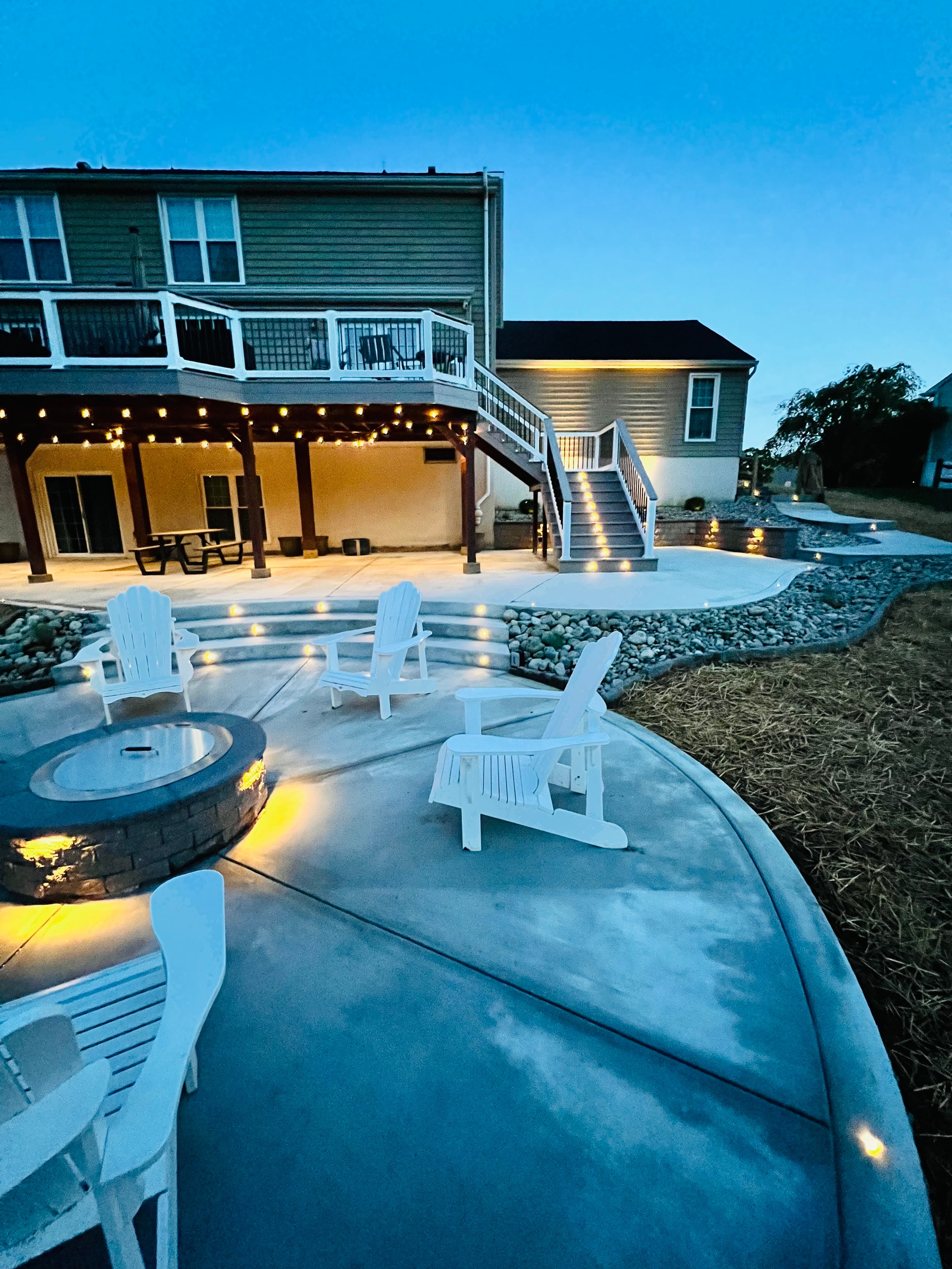 Concrete Jungle — ground-level dusk shot of circular fire pit with white Adirondack chairs, LED step lights, string lights under covered patio, and multi-level deck staircase