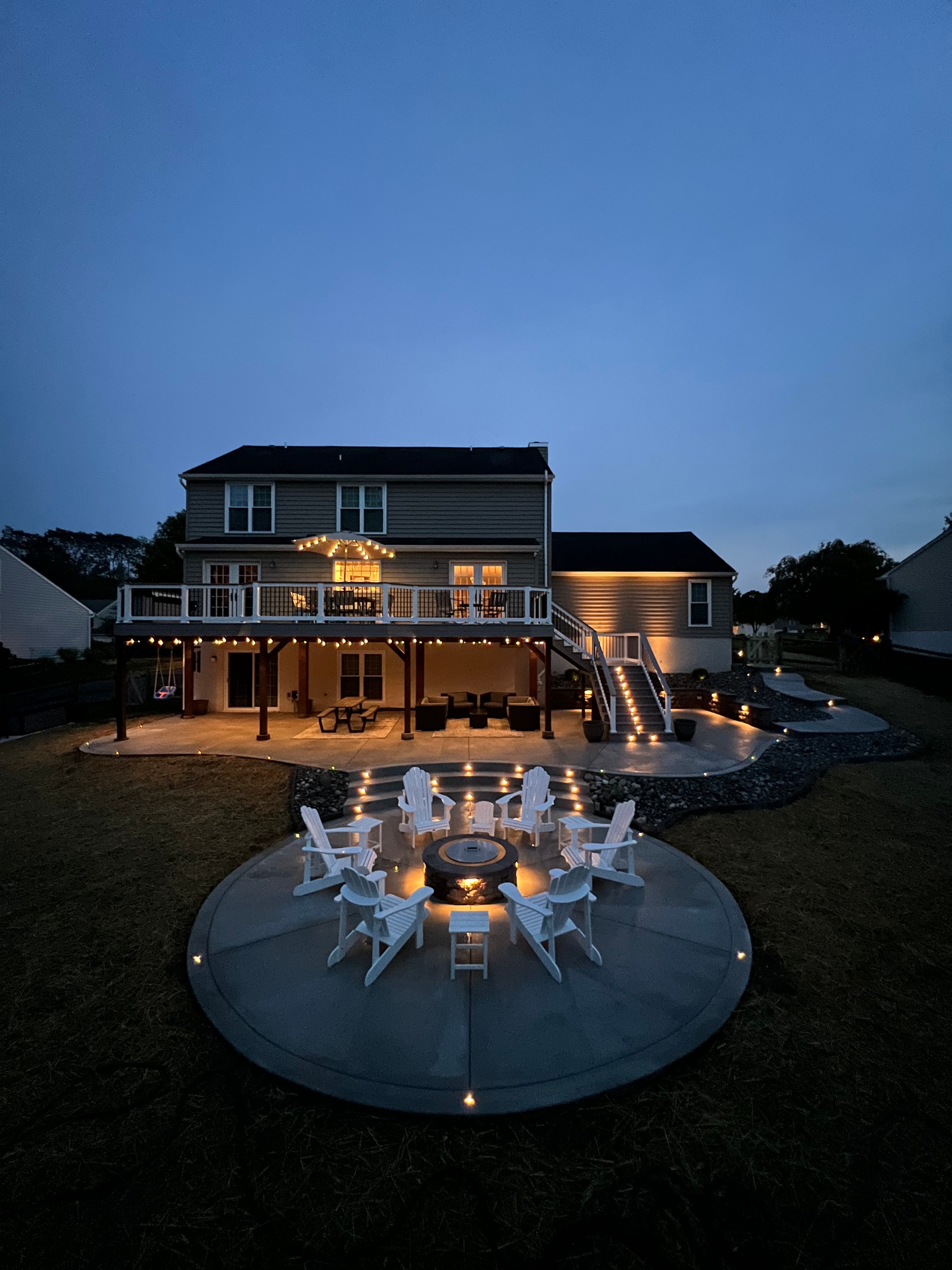 Concrete Jungle — wide dusk shot showing circular concrete fire pit pad with white Adirondack chairs, string lights, and multi-level deck glowing against blue evening sky