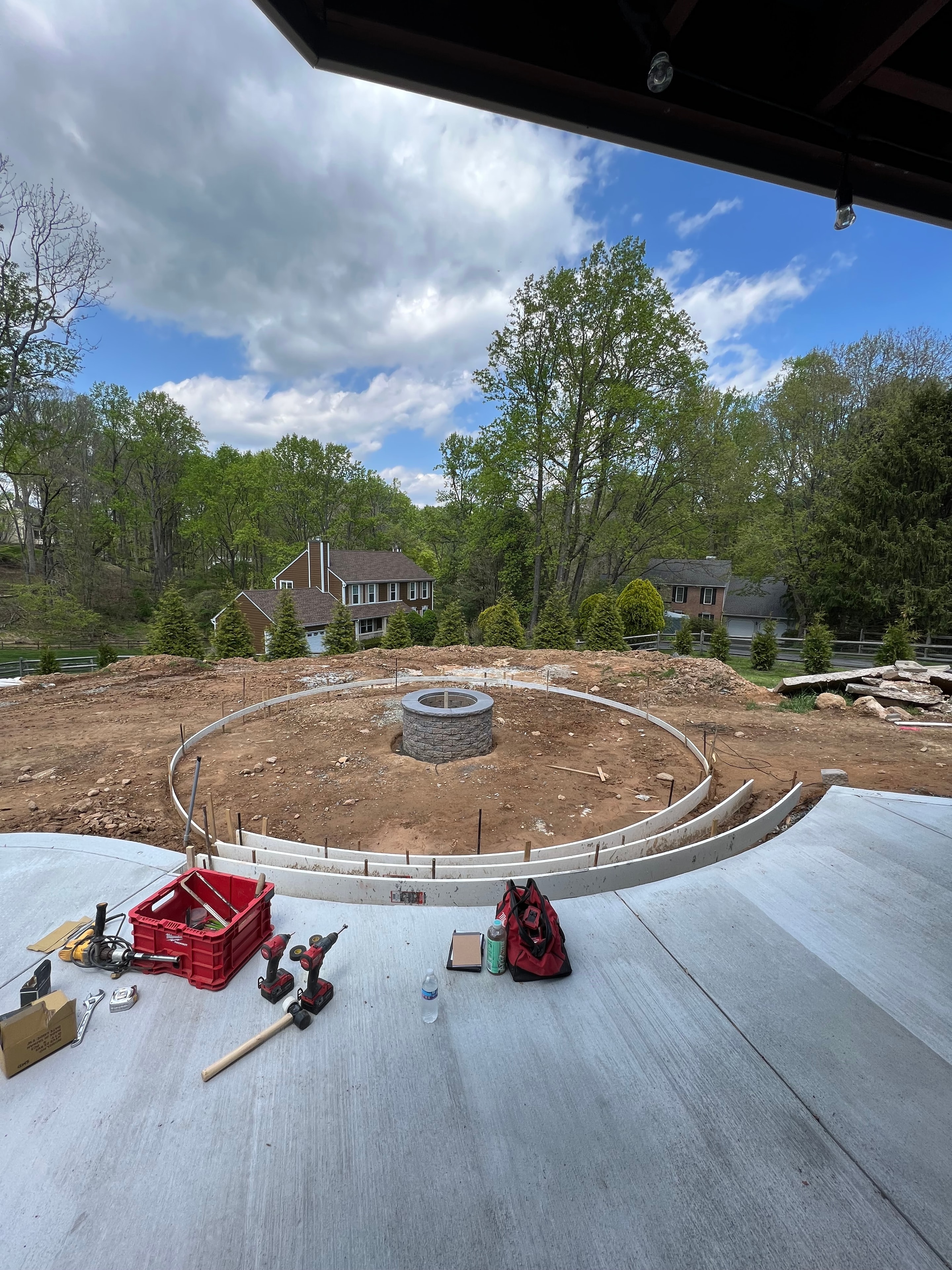 Under construction — circular fire pit form laid out on bare ground with fresh concrete patio poured, tools and red Milwaukee bag on site, blue sky with clouds
