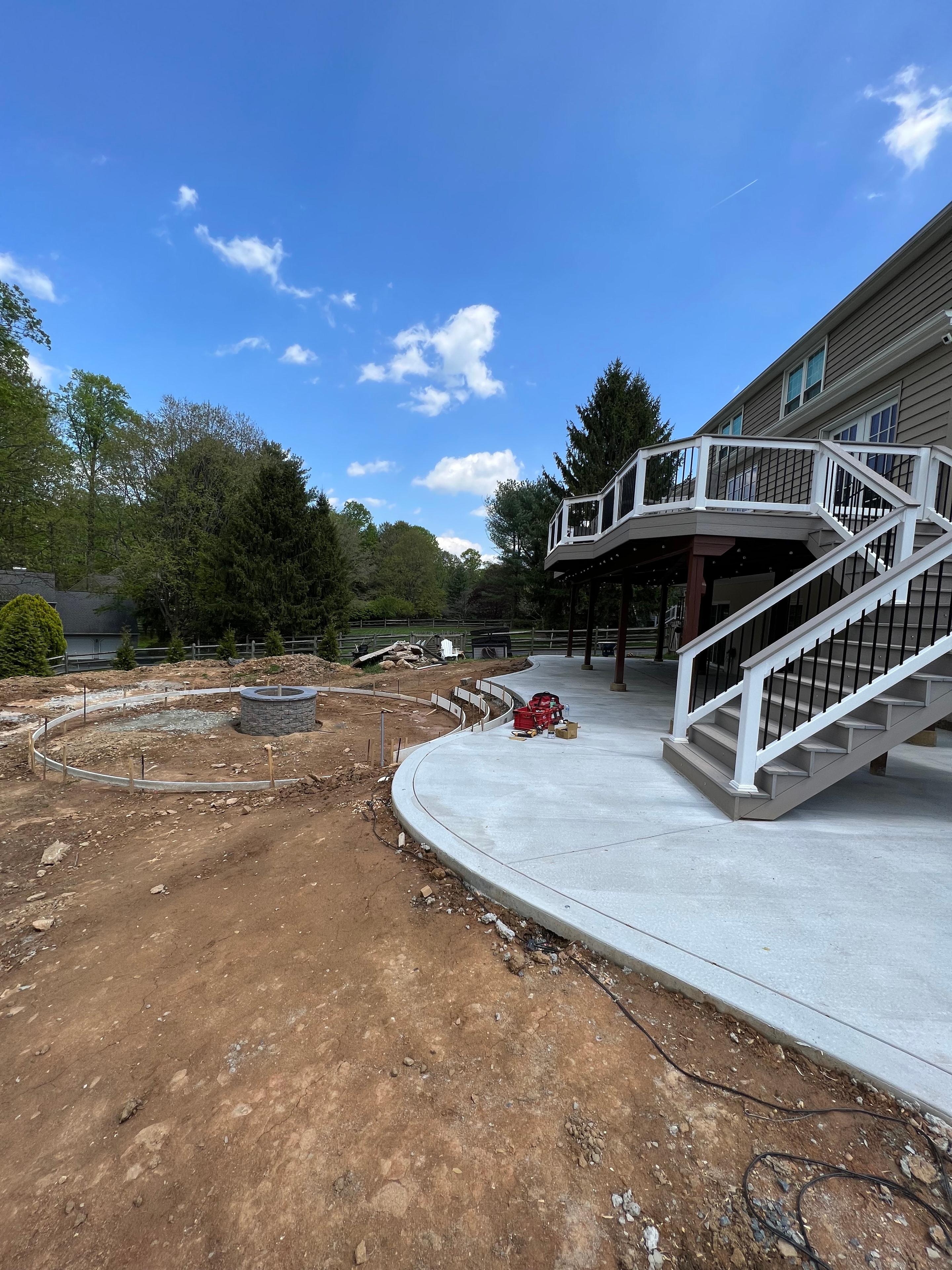 Under construction — fresh concrete patio beside house with circular fire pit form, multi-level deck framing visible, red pump on site, bare dirt surrounding