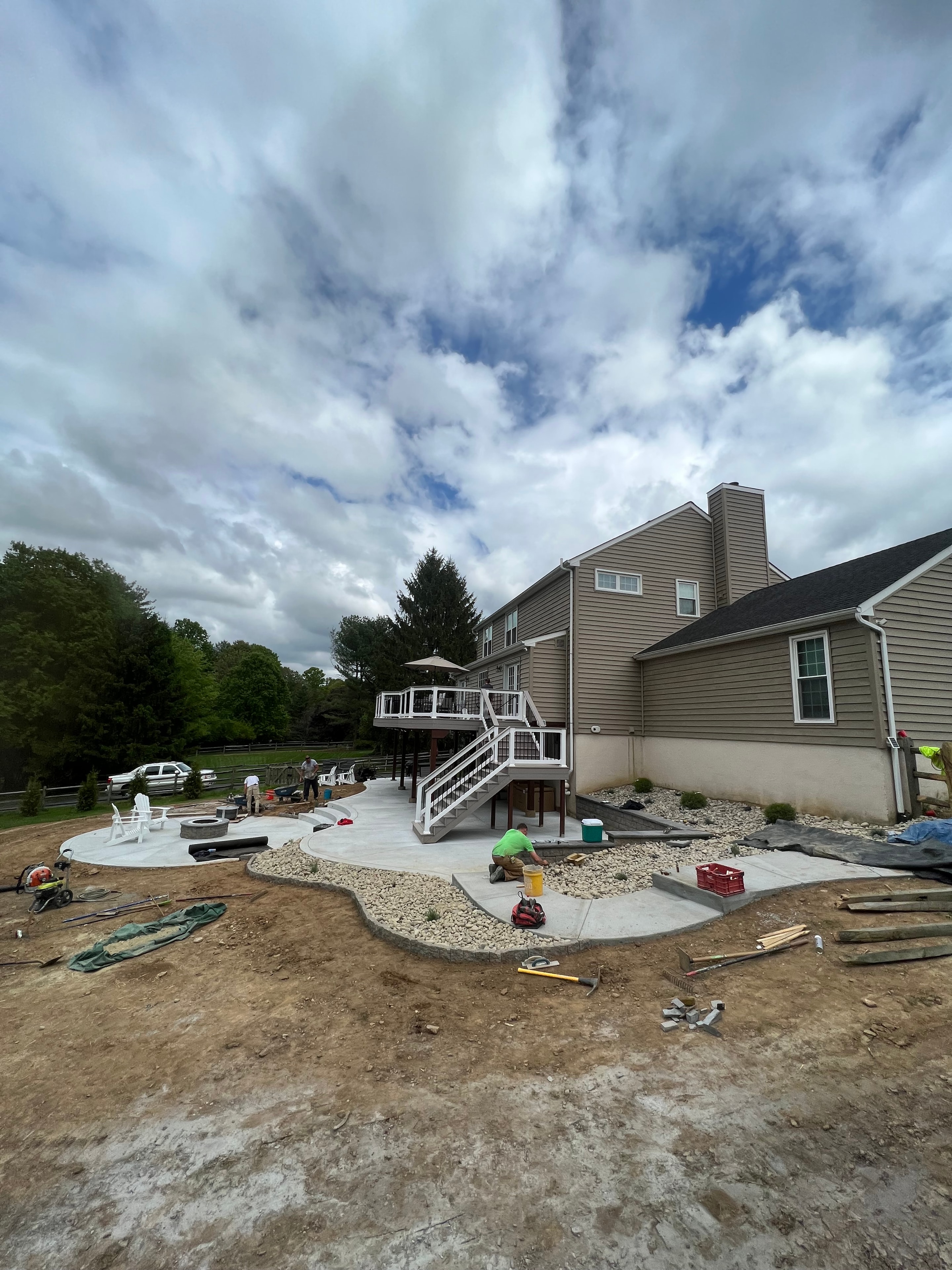 Under construction — wide shot of concrete patio with river rock border taking shape, crew working near deck staircase, bare dirt yard, white Adirondack chairs staged in background