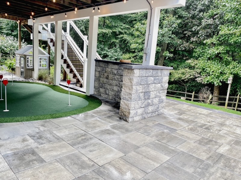 Golfing Paradise — wide daytime shot of paver patio under pergola with putting green, granite column, and string lights