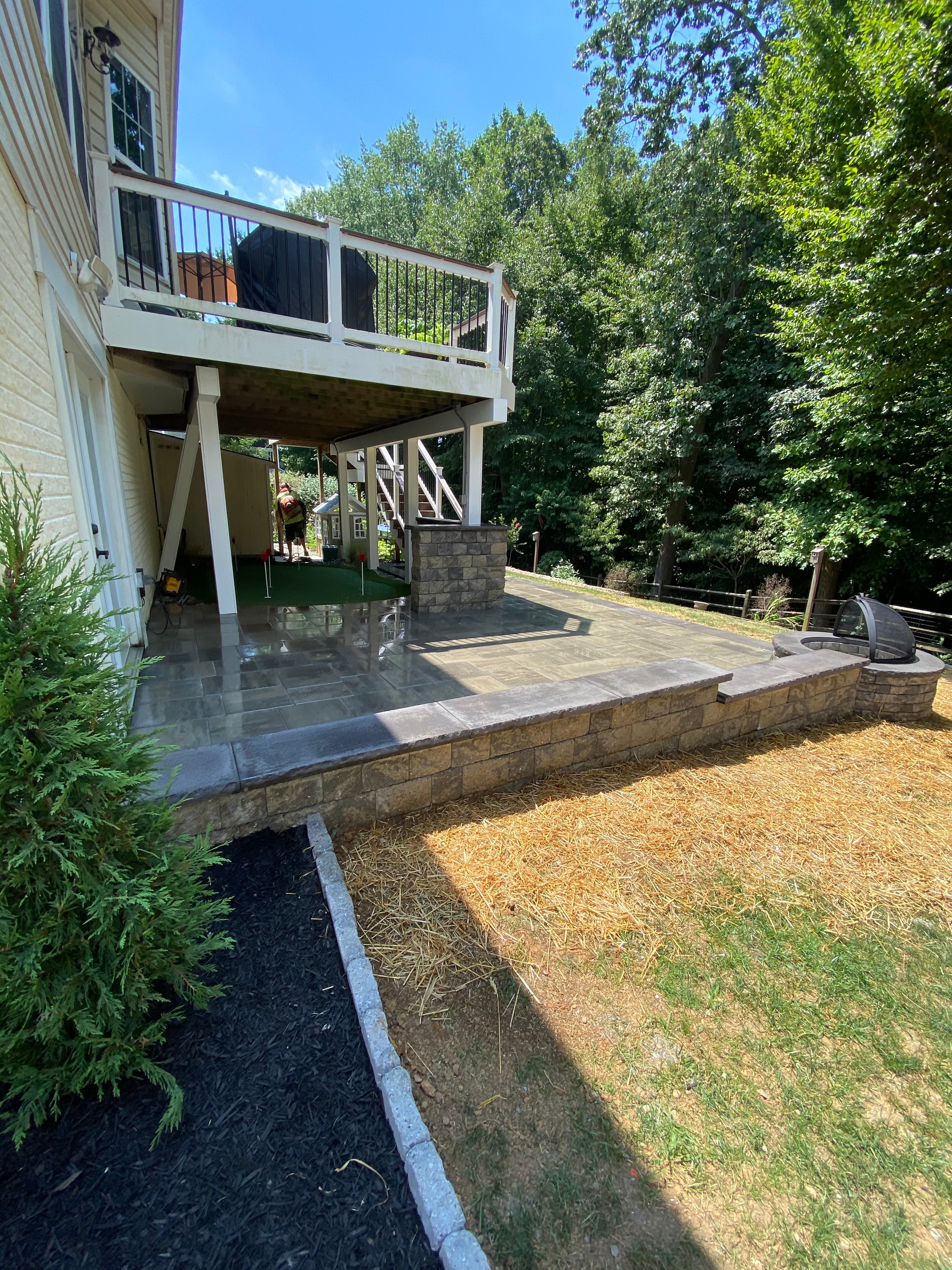 Golfing Paradise — daytime view of paver patio with pergola, granite column, putting green, and landscaping under blue sky