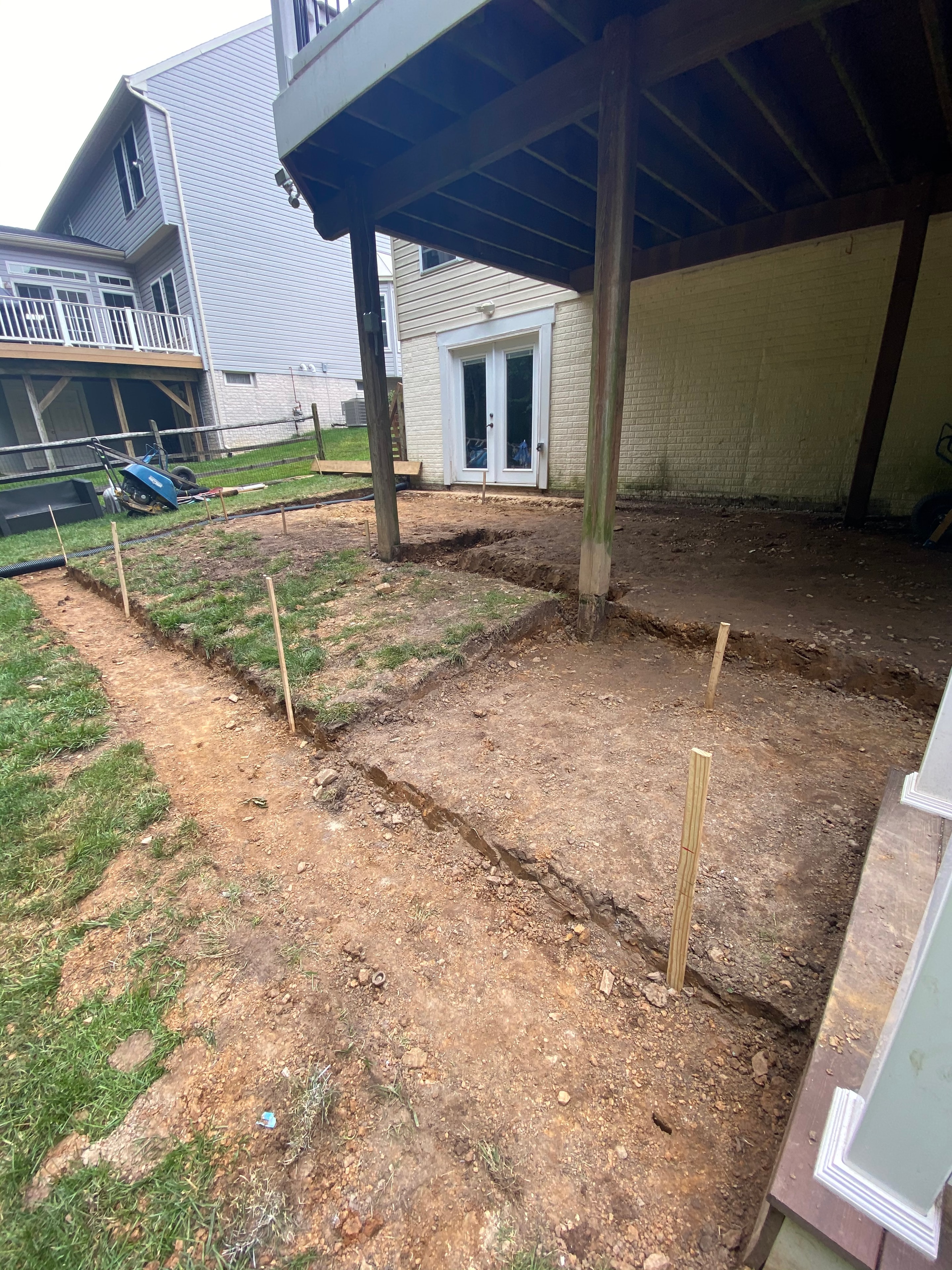 Under construction — excavated backyard with layout stakes and deck posts beside house