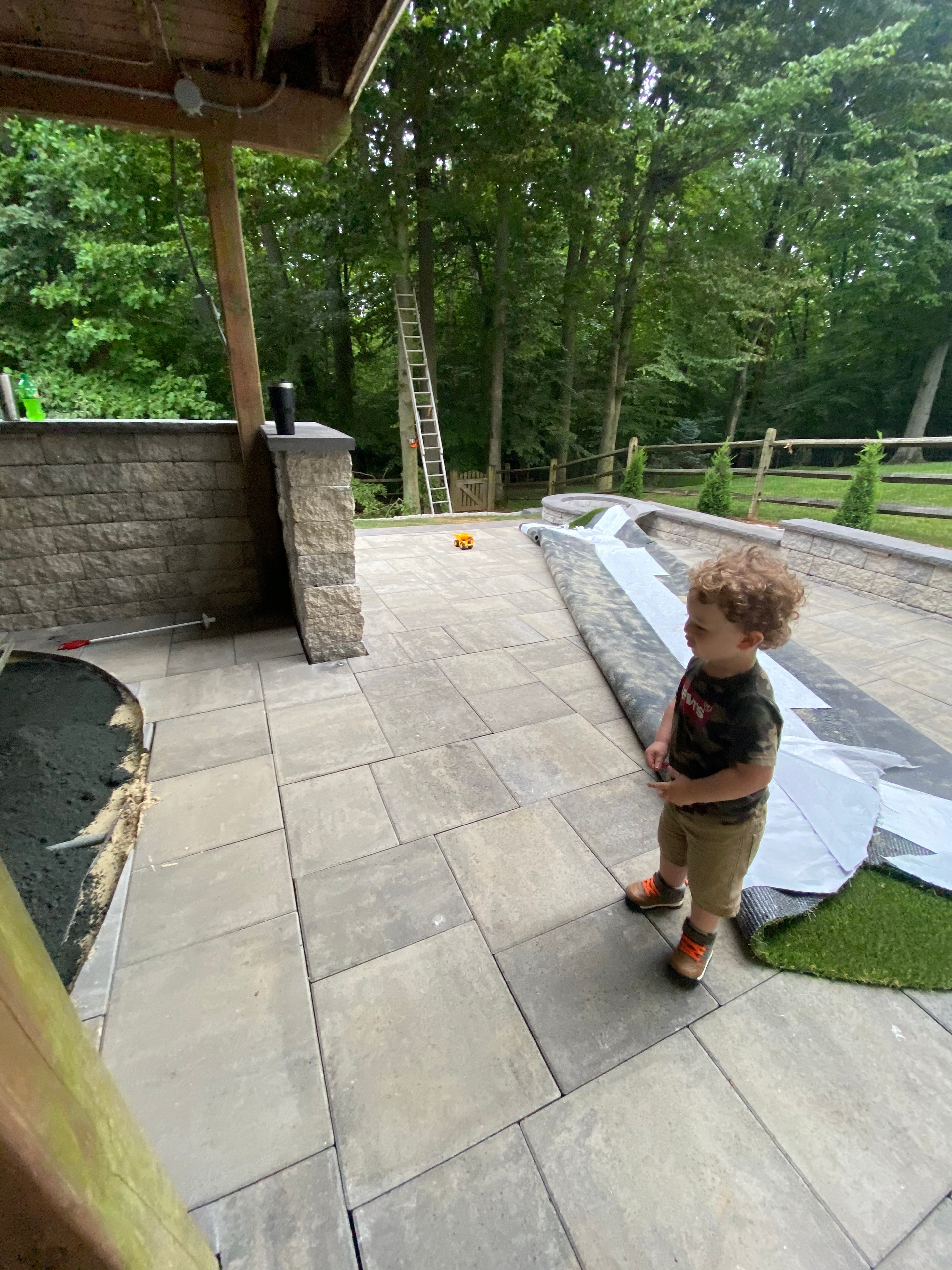 Under construction — young child walking on freshly laid paver patio with putting green turf being installed in background
