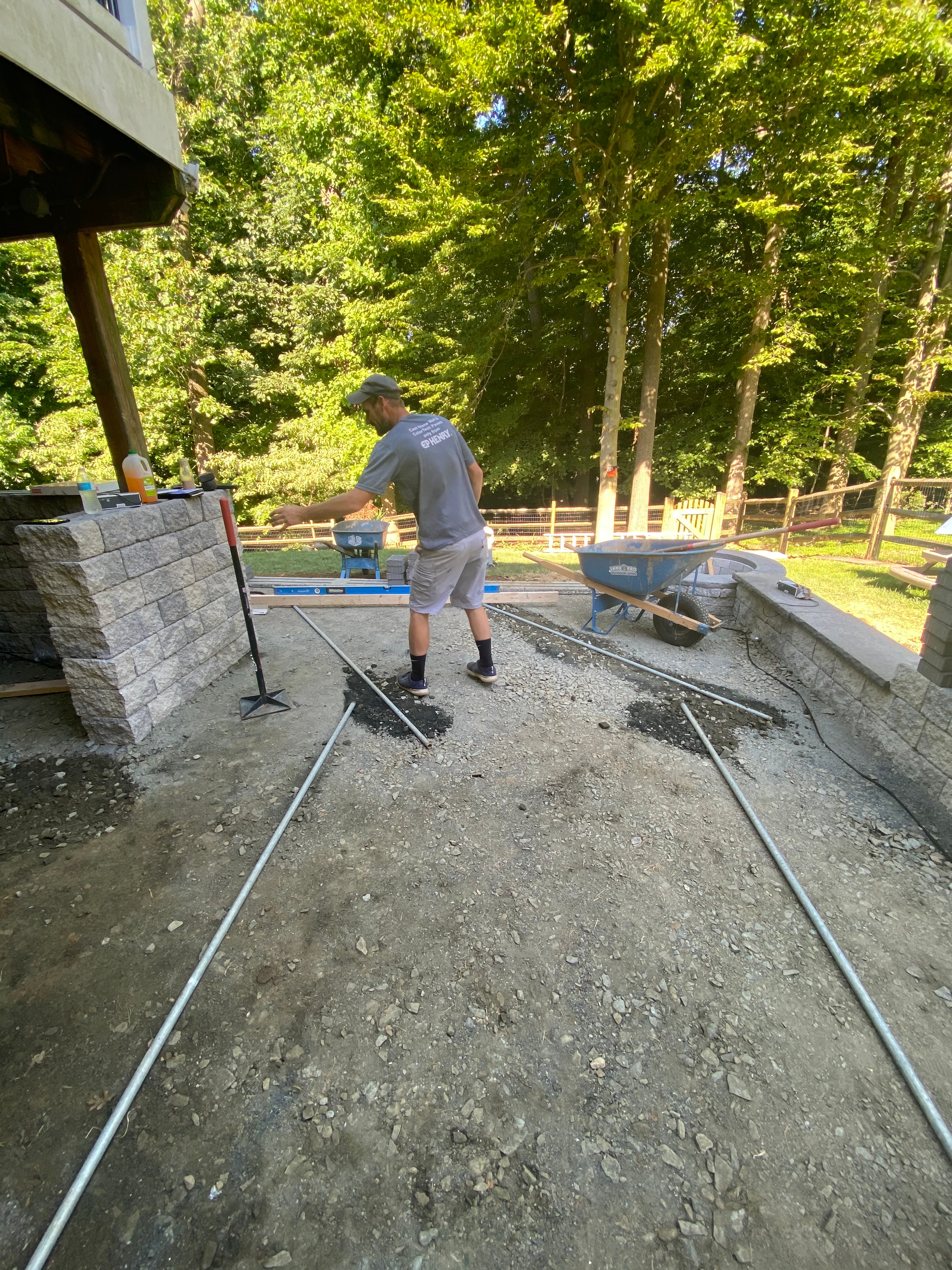 Under construction — crew member raking base material on site with granite sitting wall and pergola posts visible