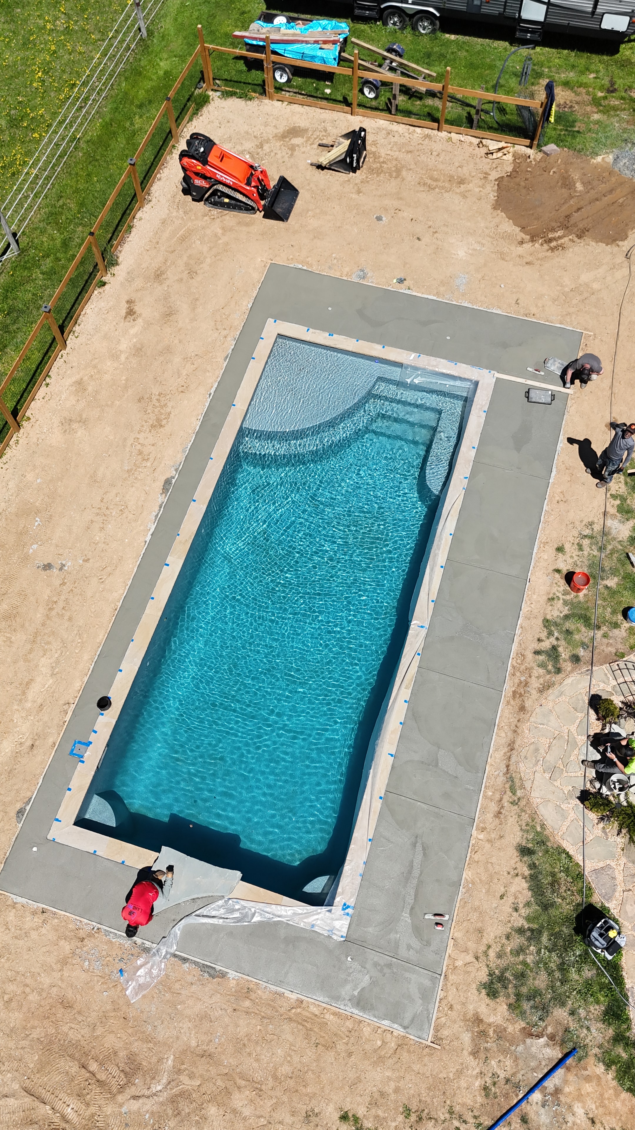 Under construction — aerial view of pool shell installed with concrete coping and crew working on surround