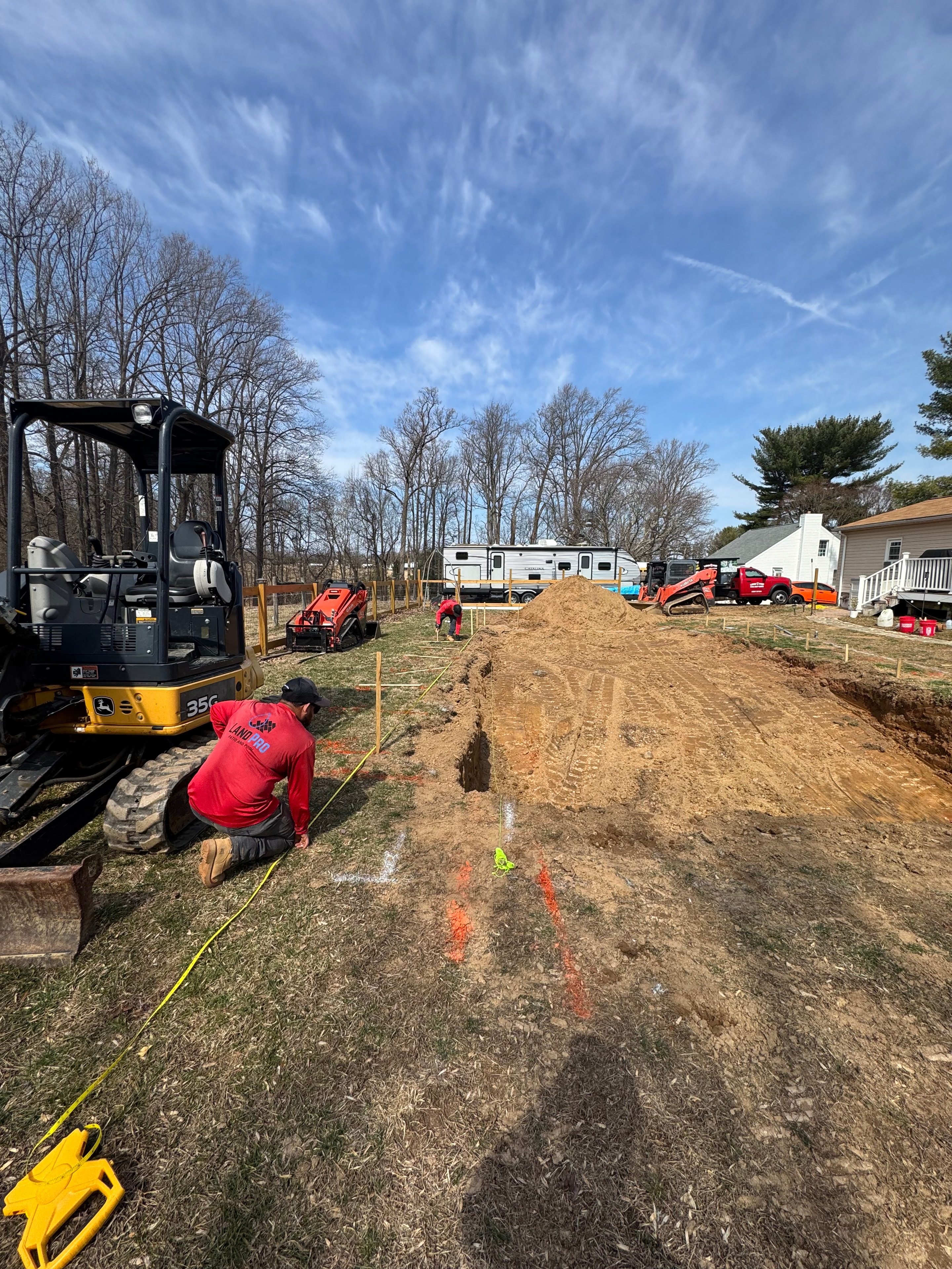 Under construction — LandPro crew member on site with excavator and equipment staged, pool area staked out