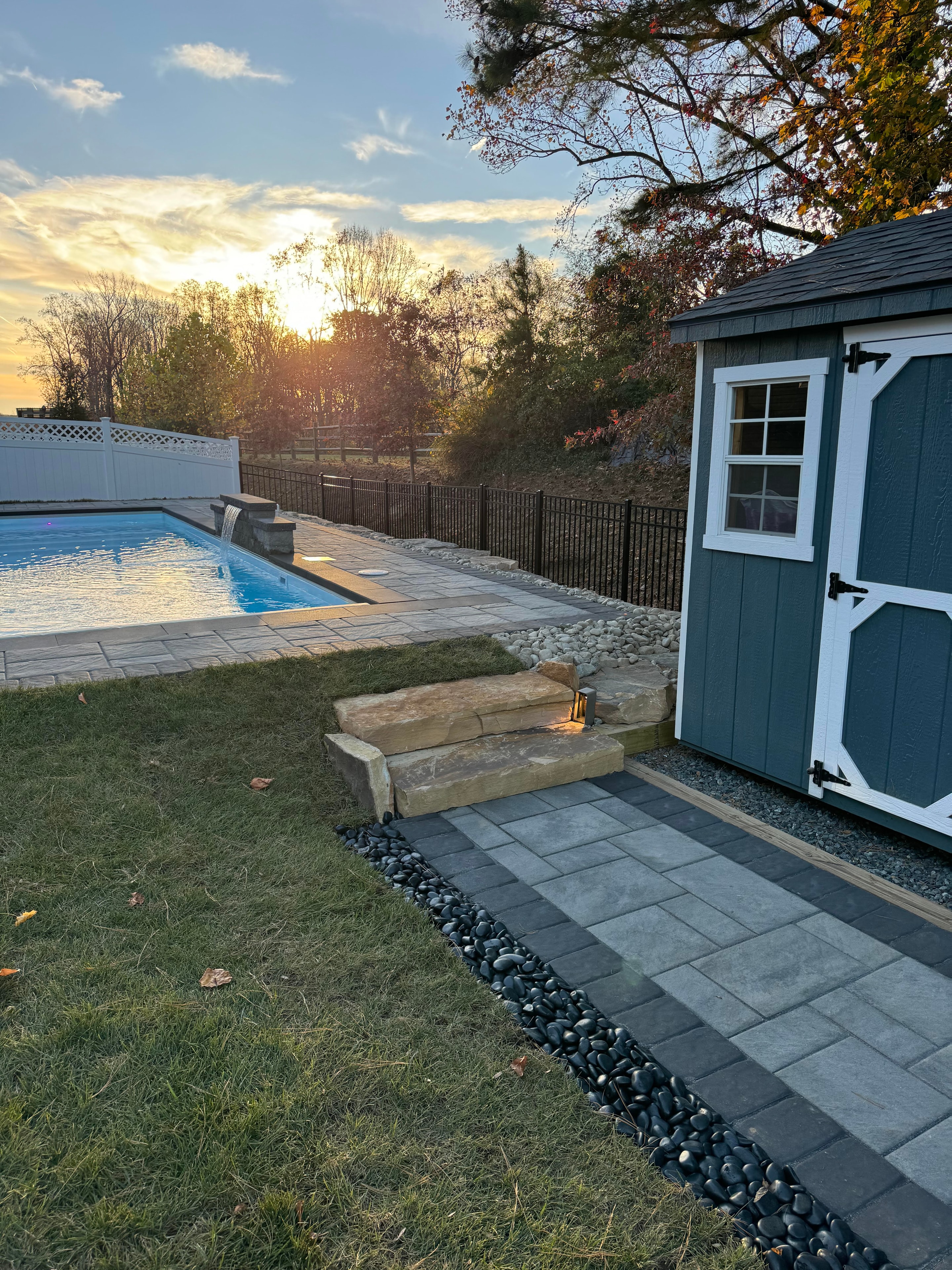 Small but Mighty — sunset view of paver pathway, natural stone steps, river rock border, and shed beside pool
