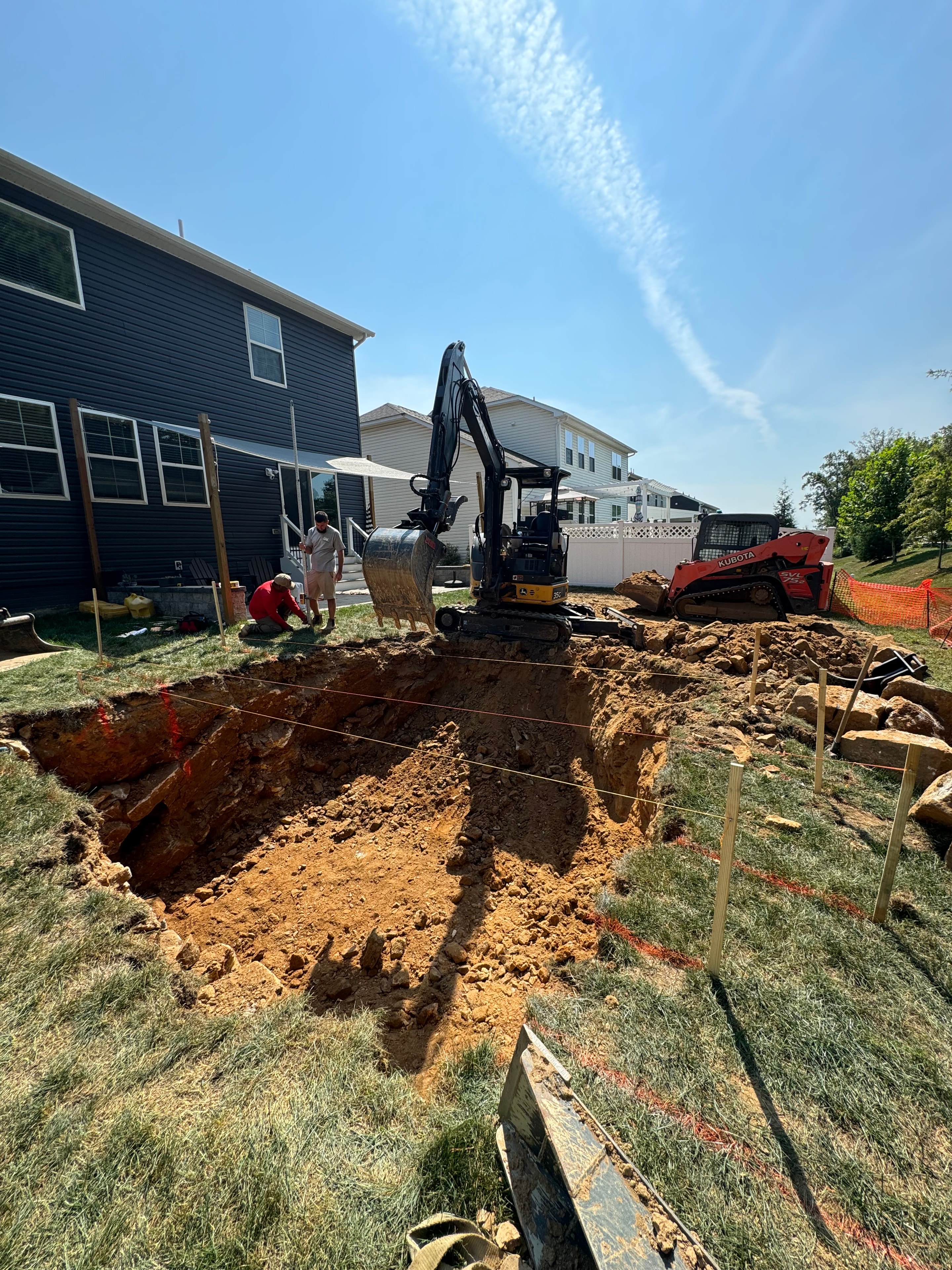 Under construction — Kubota excavator digging pool hole with crew members on site and orange safety fencing