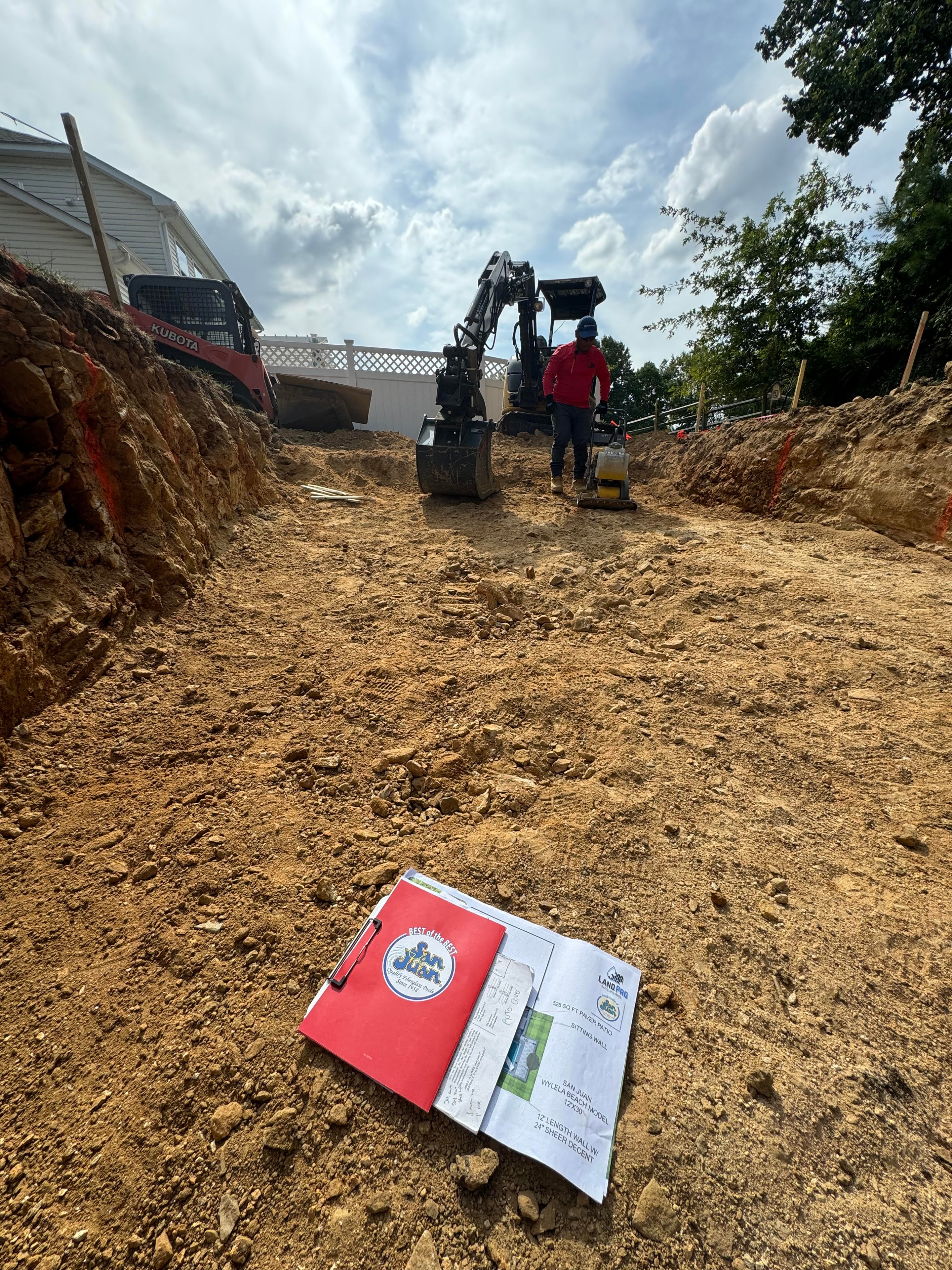 Under construction — LandPro crew member operating excavator in deep cut with San Juan site plan clipboard in foreground