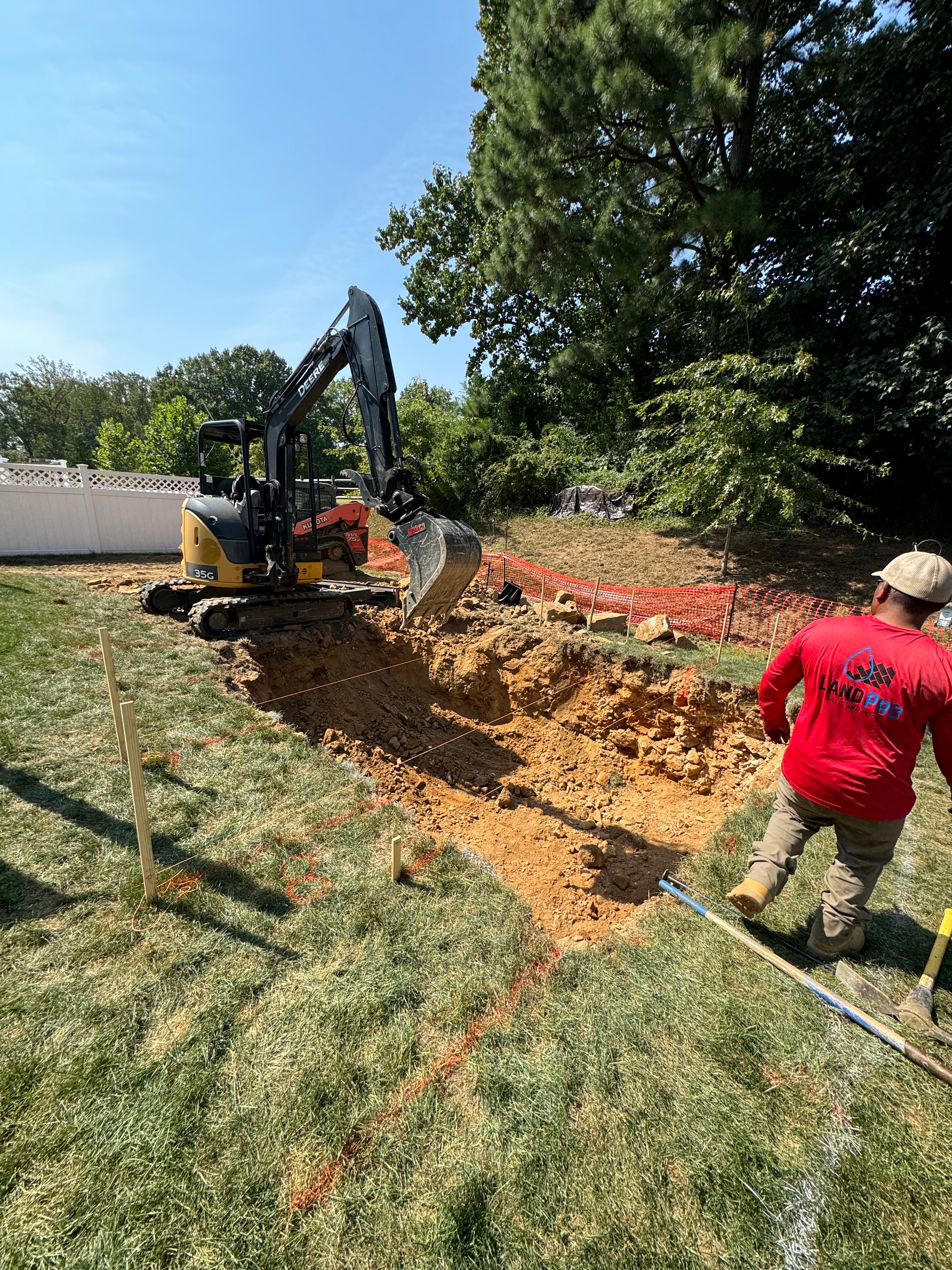 Under construction — LandPro crew member in red shirt overseeing John Deere excavator digging pool area with orange safety fencing