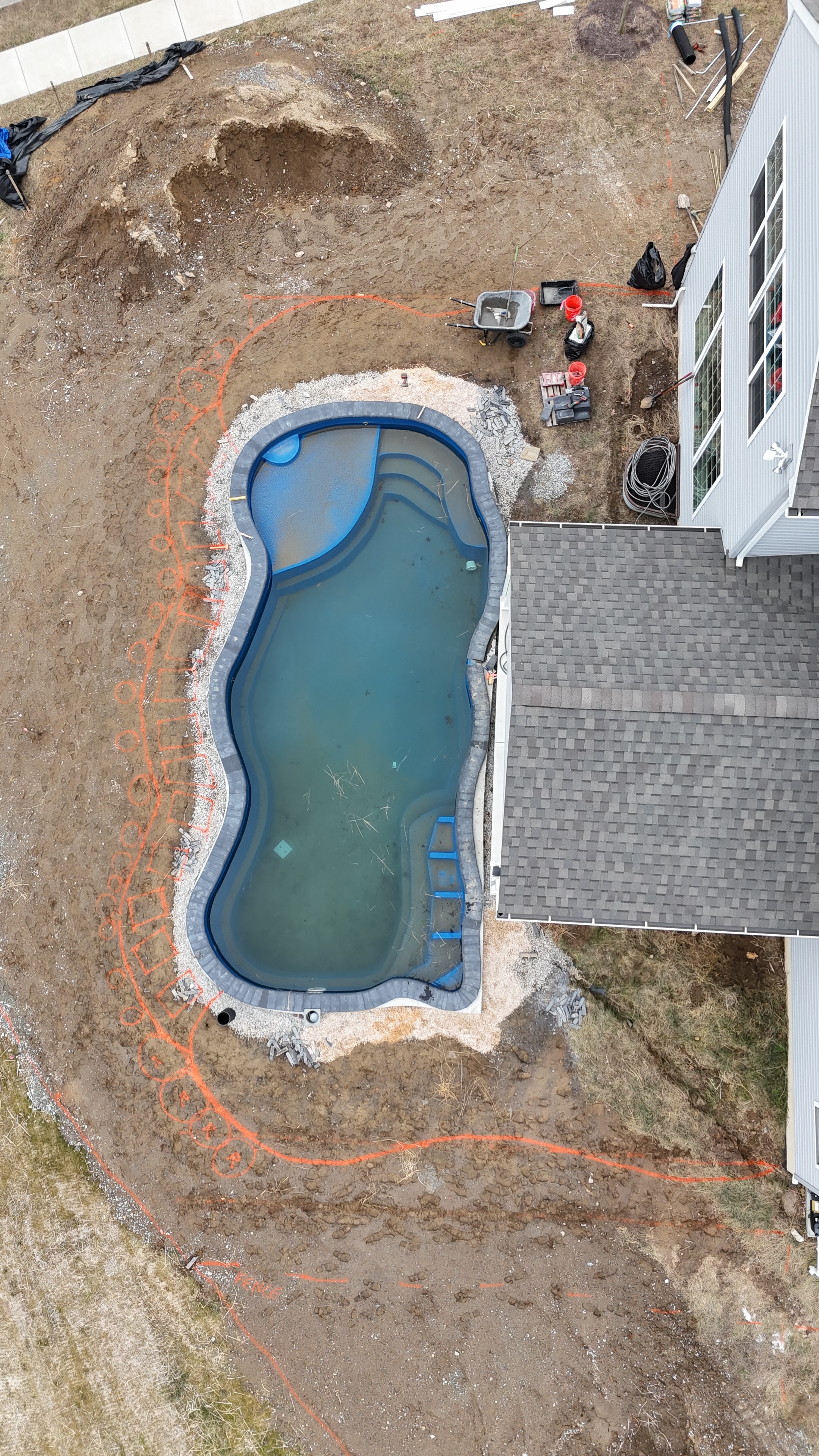 Under construction — aerial overhead shot of fiberglass pool shell set in excavated yard with orange spray-paint layout markings and solar panels visible