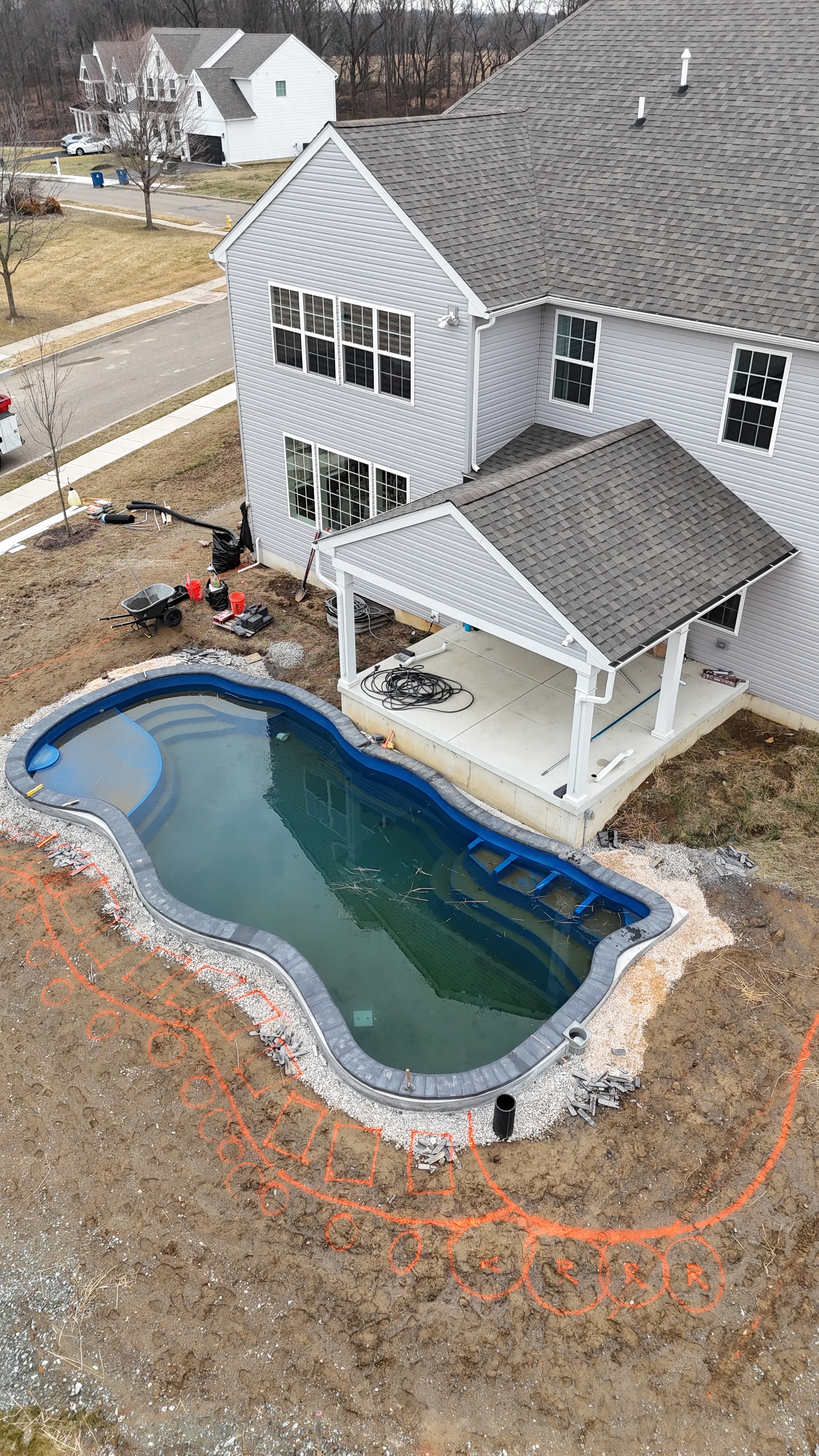 Under construction — aerial angled shot of pool shell beside house with crew working, gravel base, and orange layout markings on bare ground