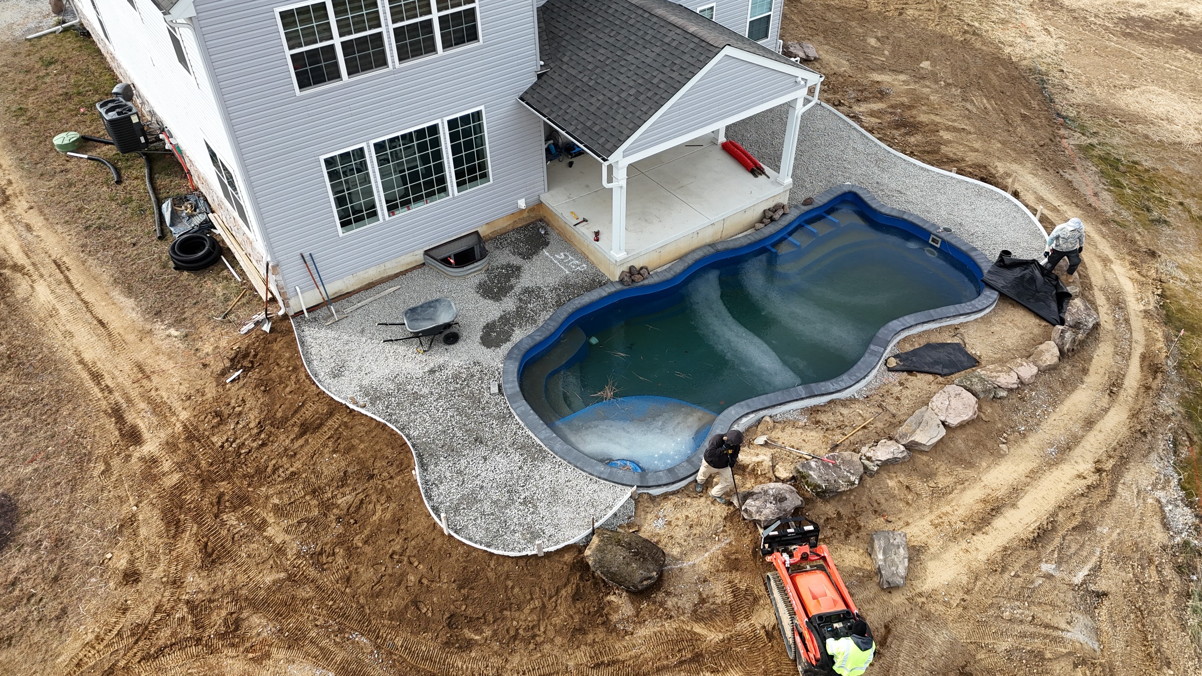 Under construction — aerial view of pool with gravel base and boulder border, crew working alongside red Kubota skid steer, concrete patio visible beside house