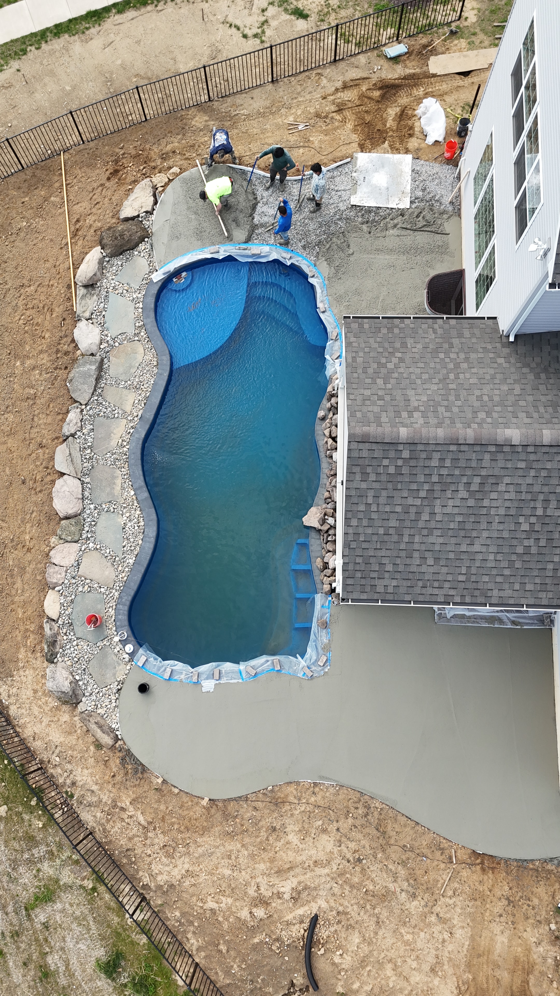 Under construction — aerial overhead shot of pool filling with water, fresh concrete patio poured on both sides, crew finishing edges, boulder border in place