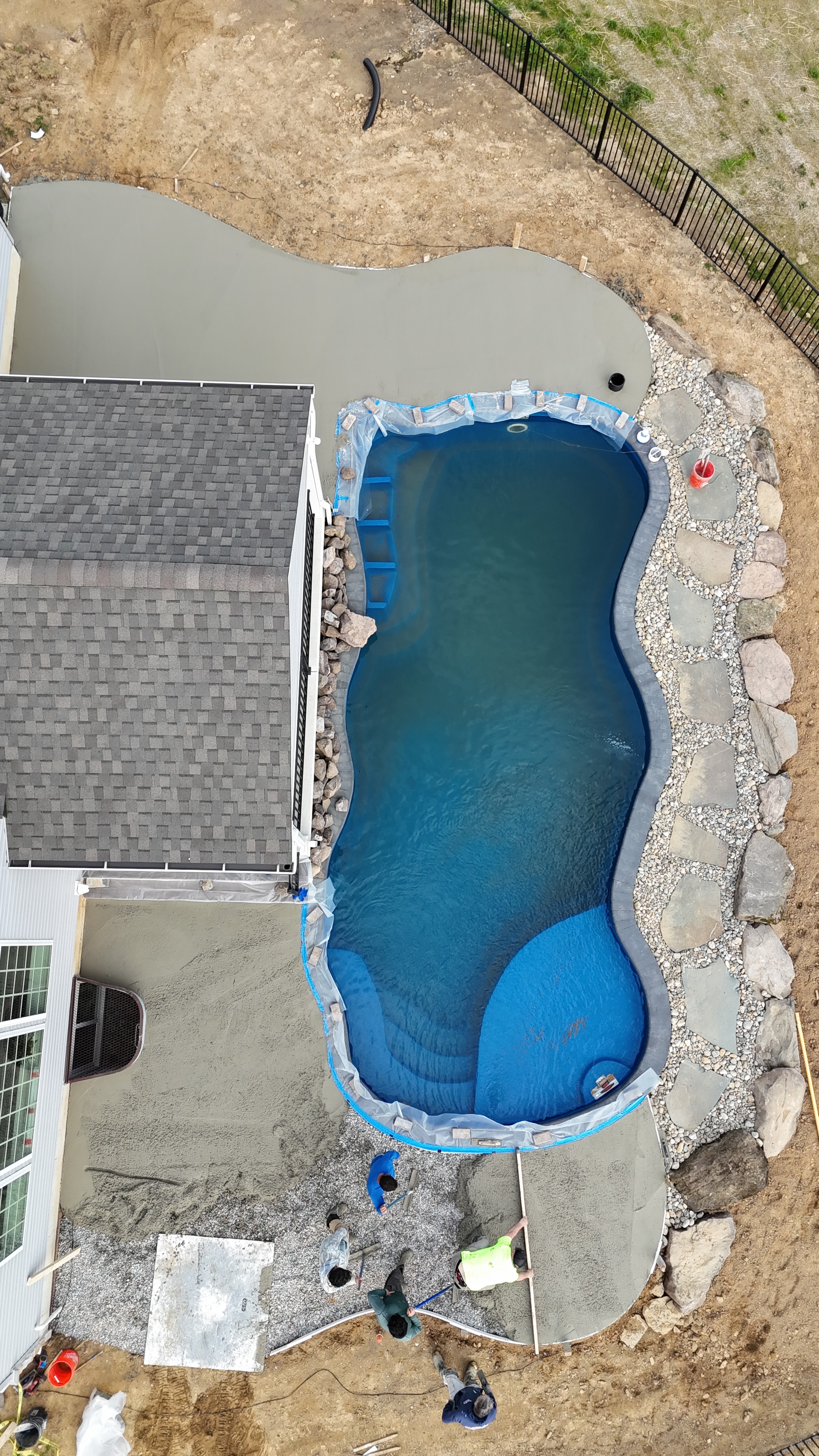 Under construction — aerial overhead shot of pool nearly full with fresh concrete patio surrounding it, crew finishing concrete work, boulder border complete